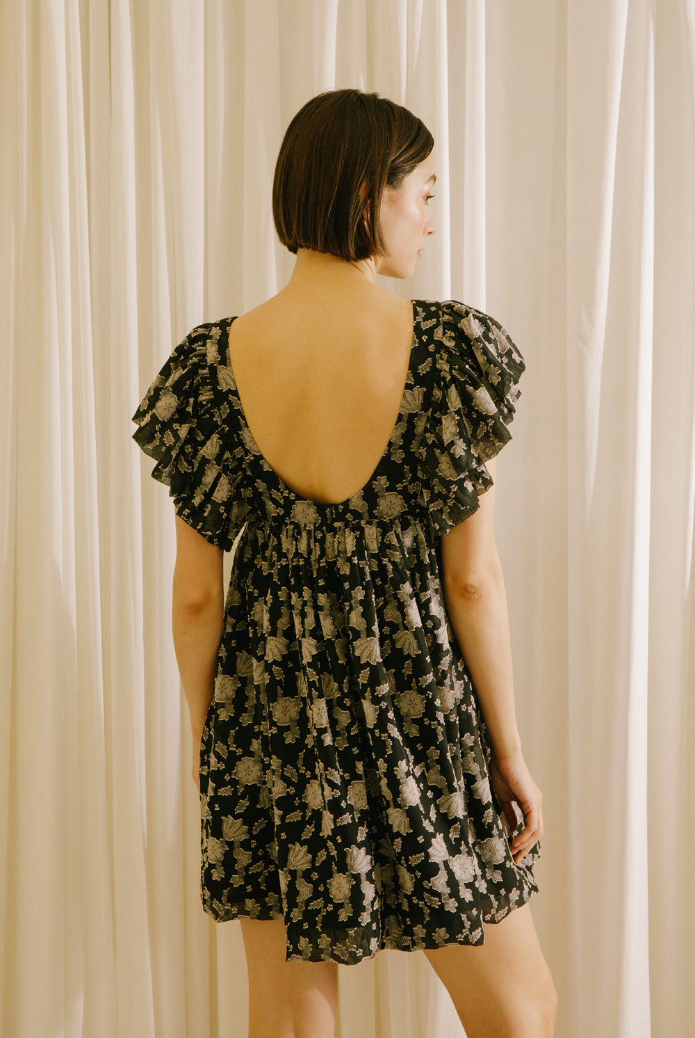 Back view of a woman wearing a black dress with a beige pattern, flutter sleeves and a v-neckline against a beige curtain background