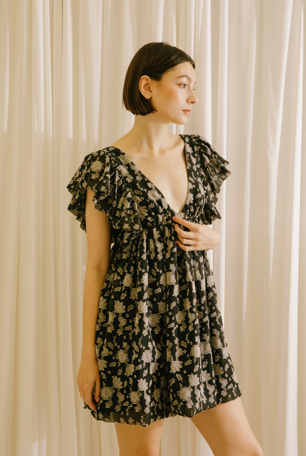 Side view of a woman wearing a black dress with a beige pattern, flutter sleeves and a v-neckline against a beige curtain background