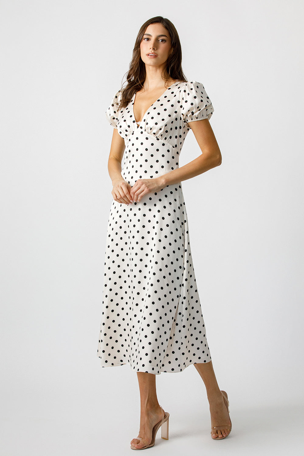 A woman wearing a white satin midi dress with ivory polka dots, short puff sleeves, and a v-neckline.