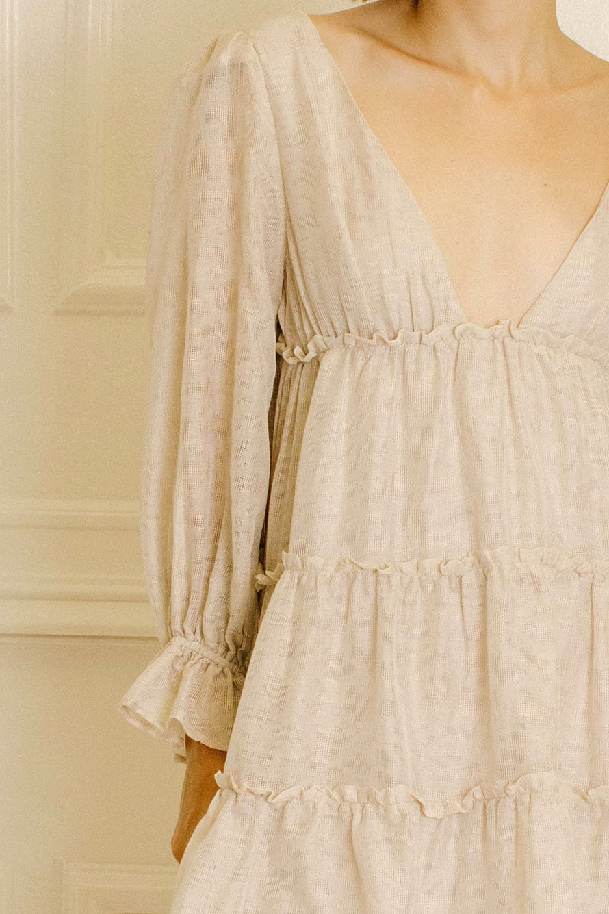 A close-up of a model wearing a light beige linen dress with a deep V-neck, gathered ruffle details, and long balloon sleeves.