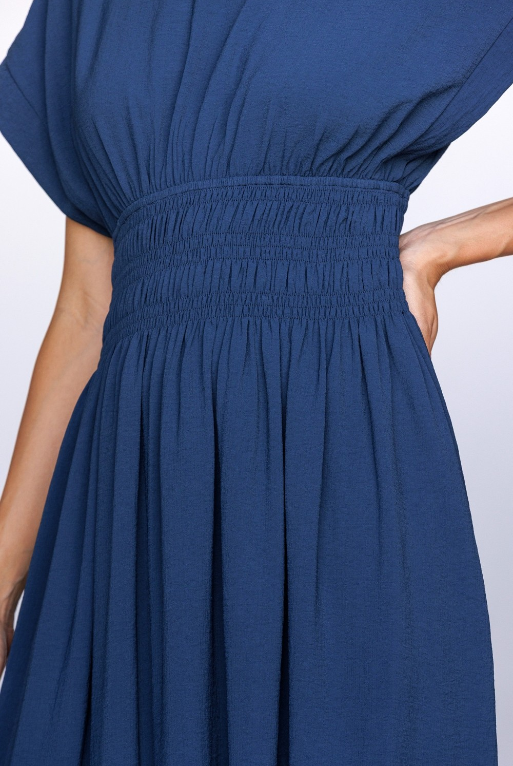 Close-up of a person wearing a navy midi dress with a smocked waist, relaxed bodice and dolman sleeves against a white wall.