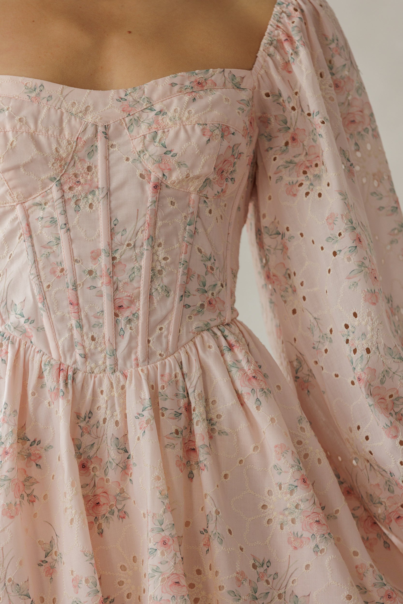 Close-up of a light blush pink eyelet dress with a delicate pink and green floral print, featuring a smocked bodice, floral embroidery and long sleeves.