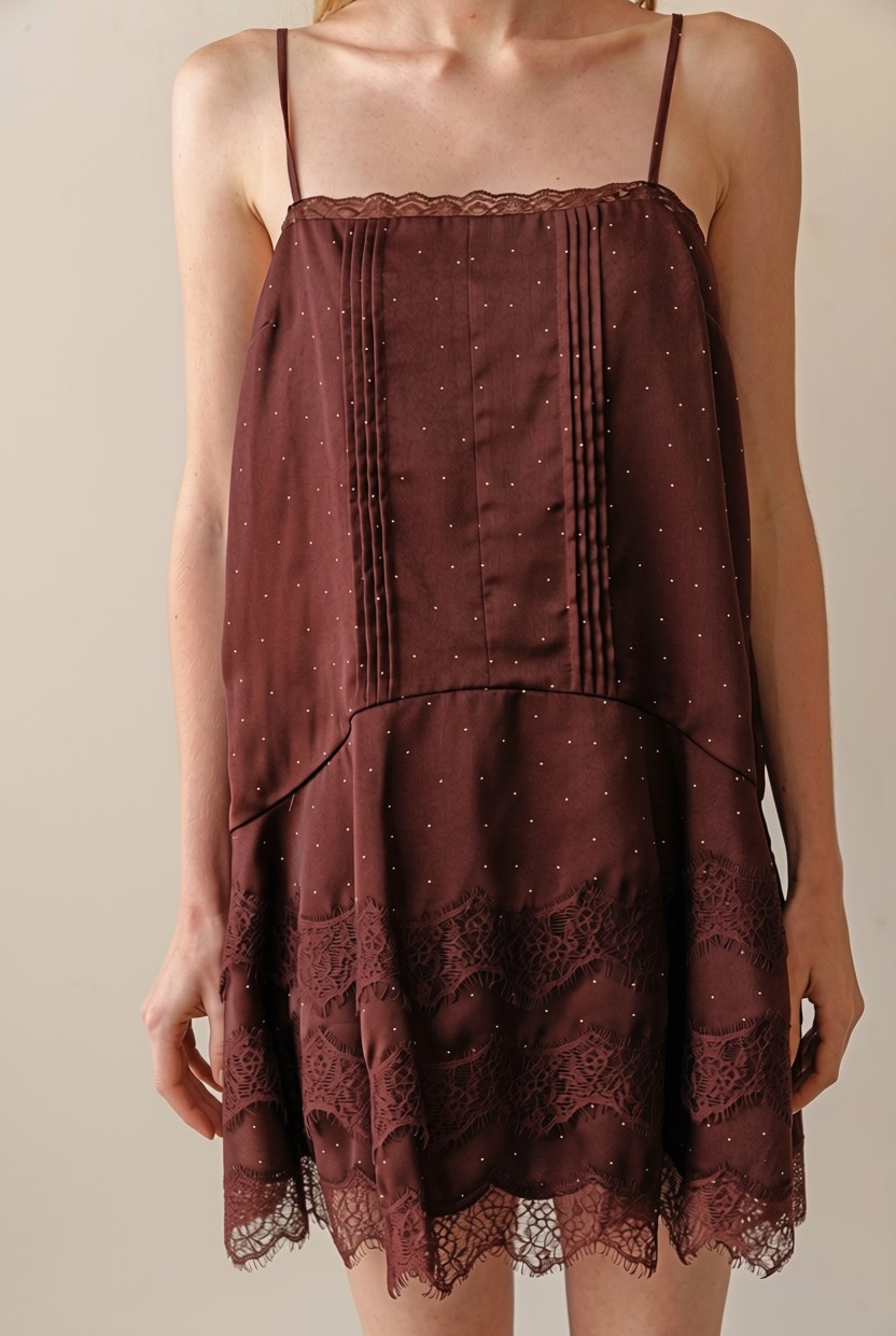 A woman wearing a dark brown spaghetti-strap mini dress with a sheer, dotted fabric and a lace-trimmed hem, shown against a plain beige background.