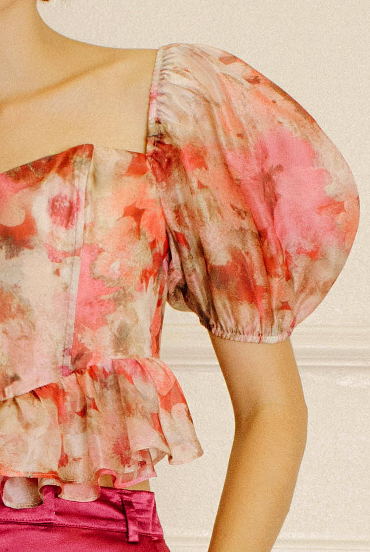 A close-up of a model wearing a sheer puff-sleeve blouse with a pink, red, and beige watercolor floral print, paired with a bright pink skirt.