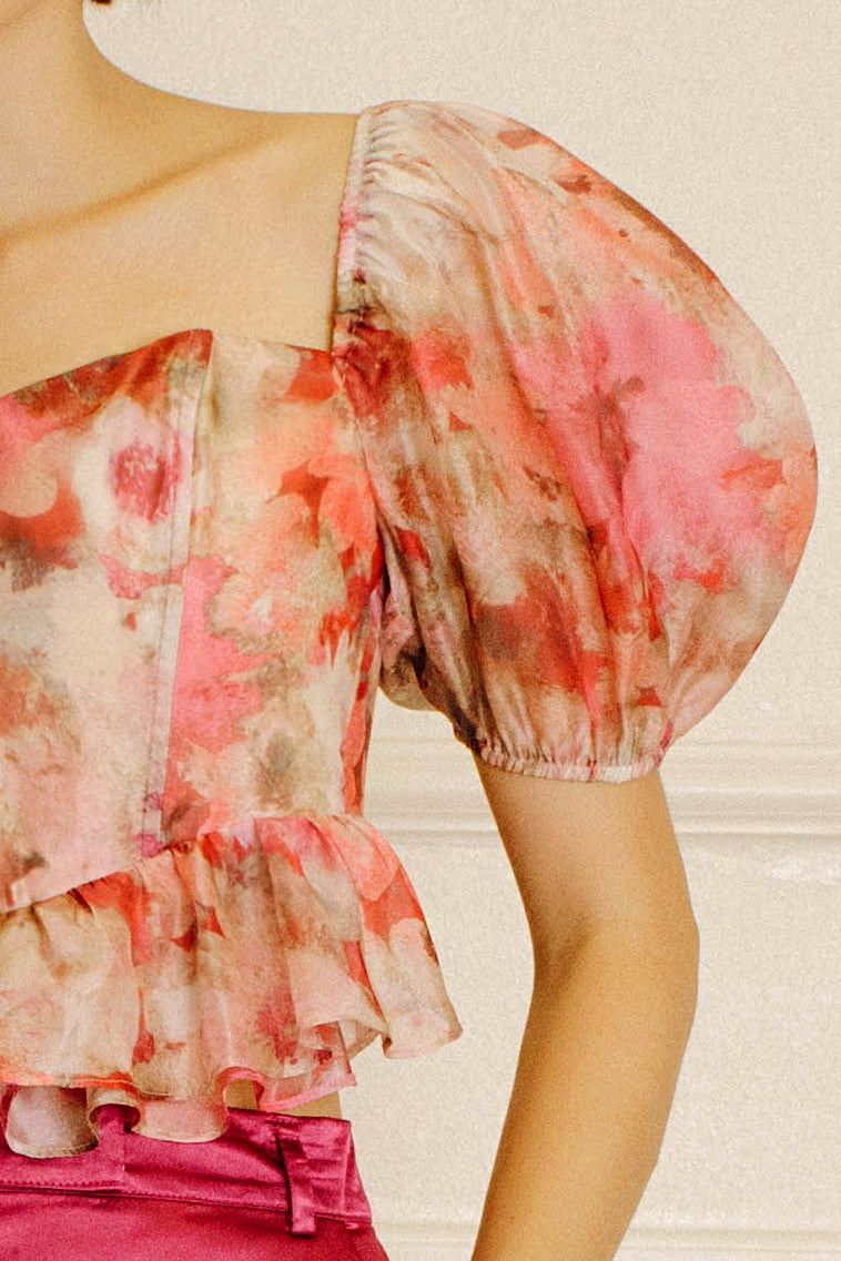 A close-up of a model wearing a sheer puff-sleeve blouse with a pink, red, and beige watercolor floral print, paired with a bright pink skirt.