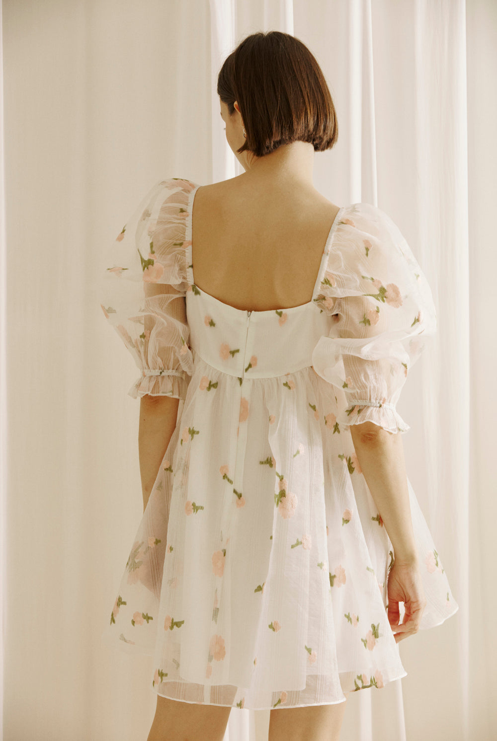 Back view of a woman wearing a white dress with a top layer of a flower embroidered and textured sheer fabric, puffy sleeves and a back invisible zipper against a light curtain background