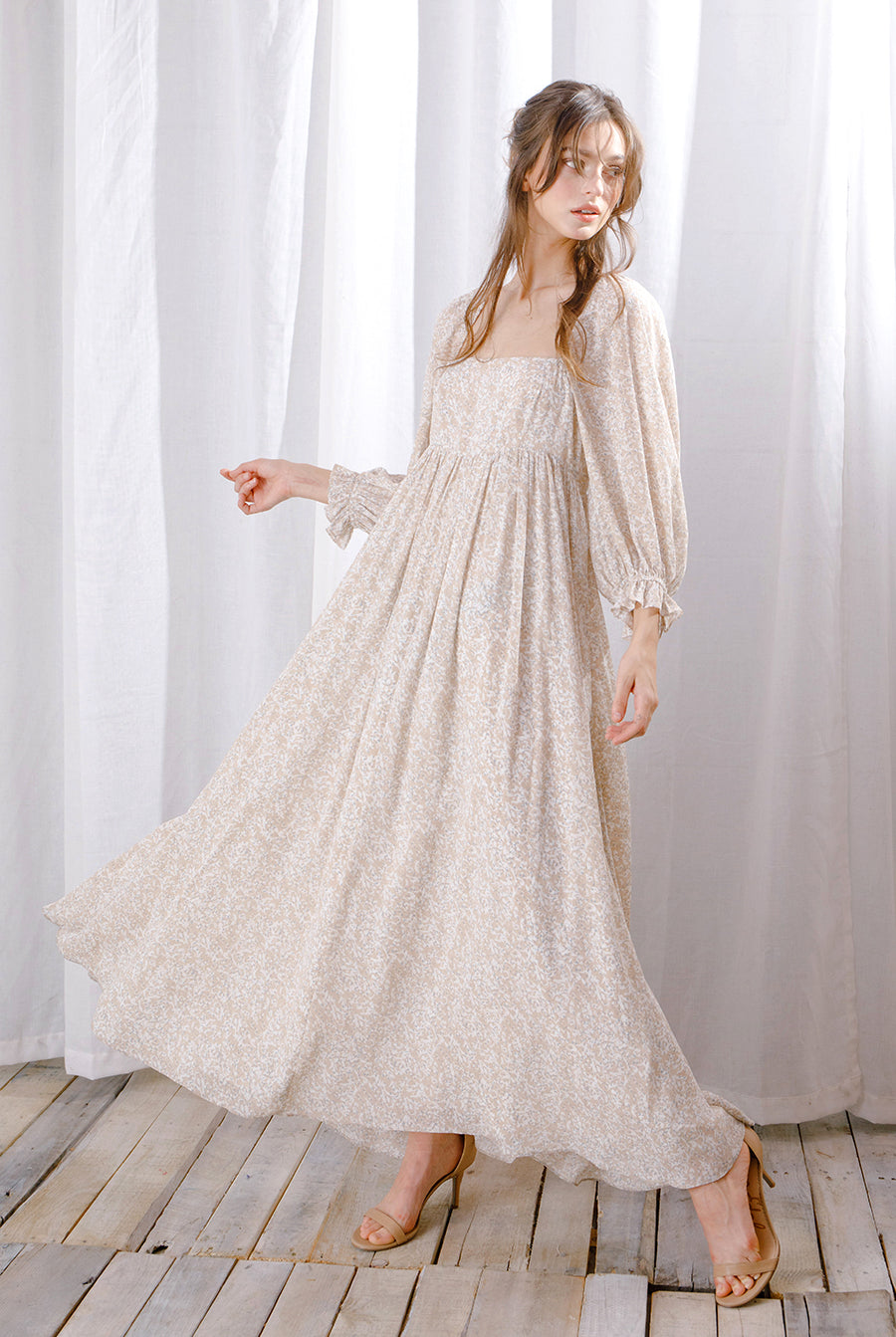 Alternative view of a model wearing a long, light-colored dress standing against a white curtain.