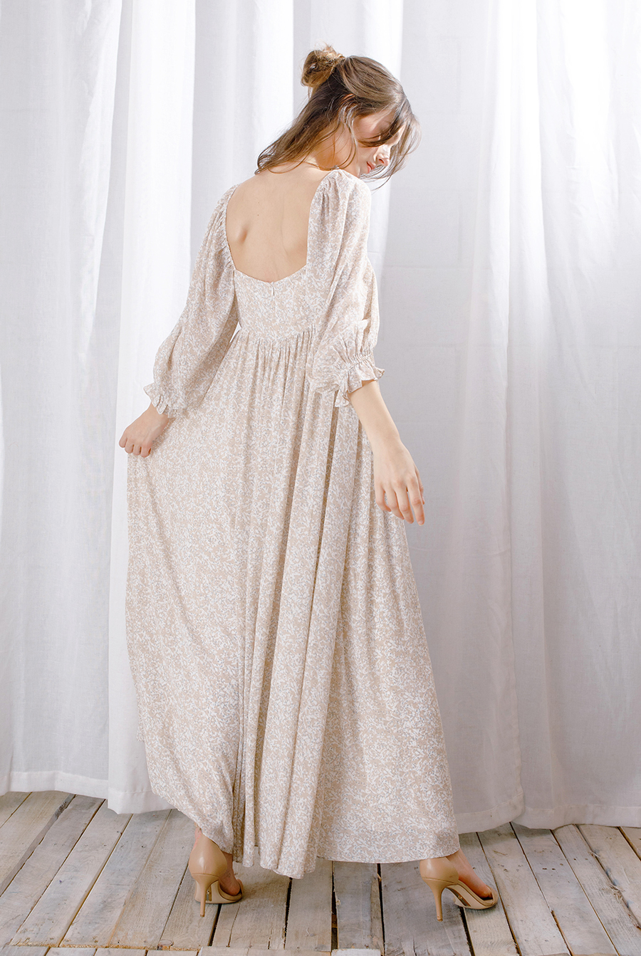 A woman wearing a long-sleeve maxi dress with a delicate beige and white floral pattern, shown from the back. She is standing on a light wooden floor in front of white sheer curtains.