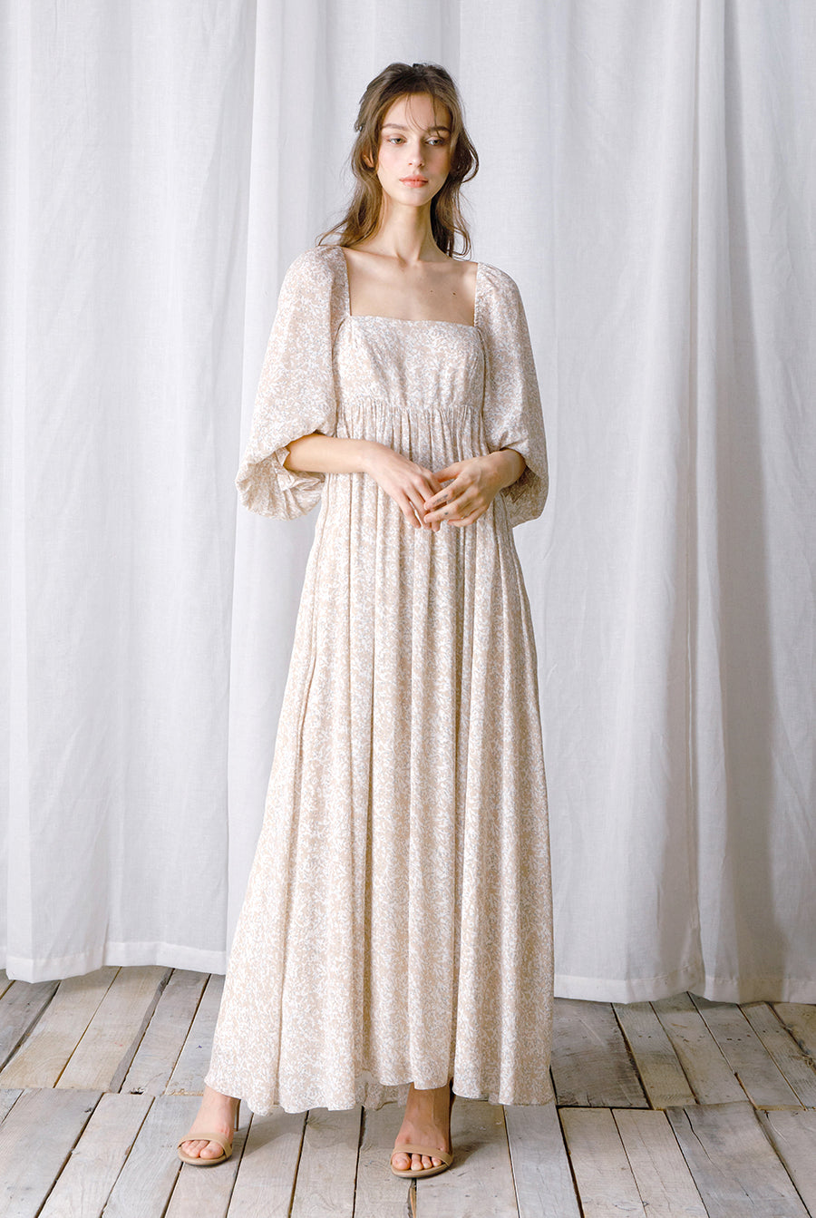 Woman wearing a long, light-colored dress standing against a white curtain.