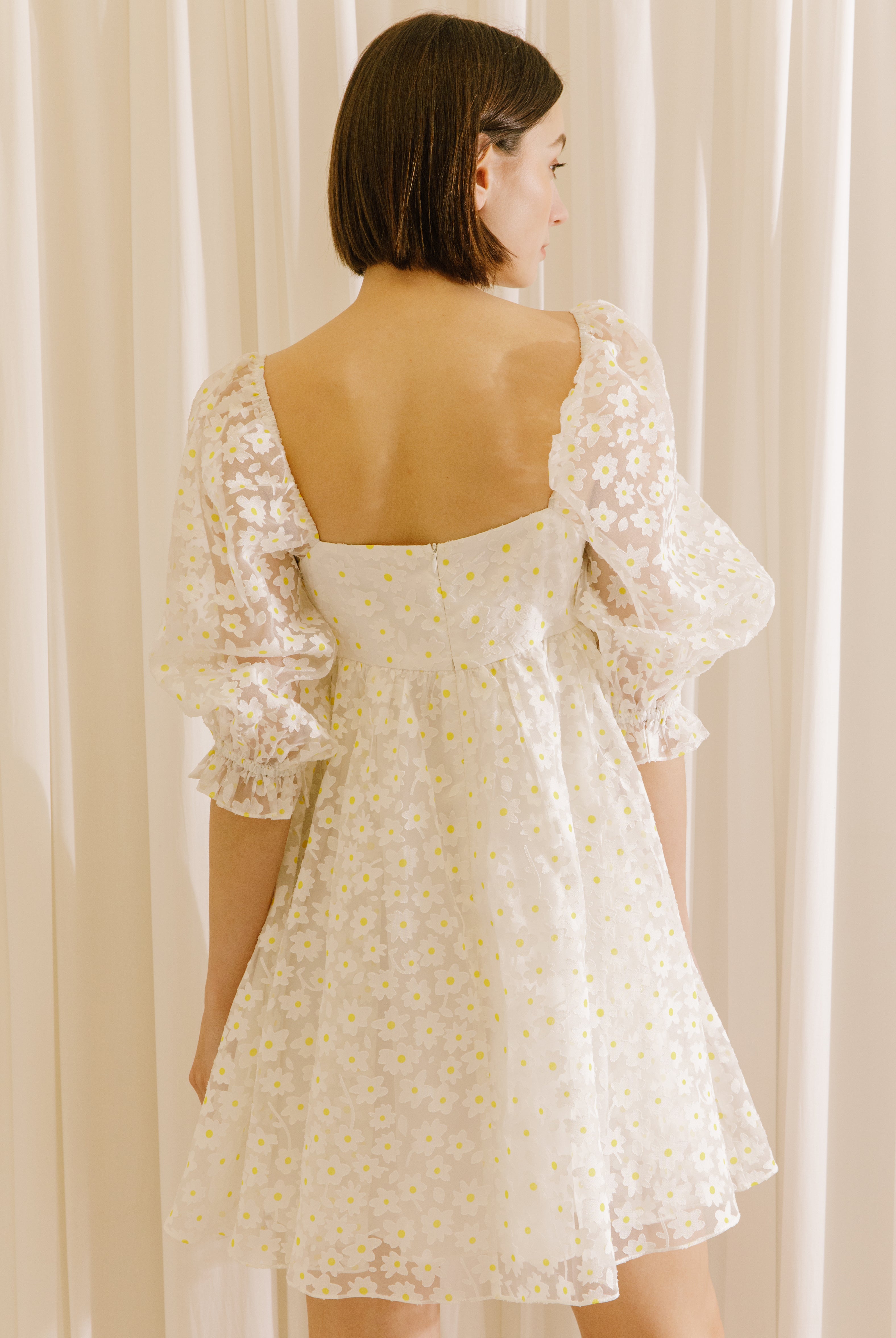Back view of a woman wearing a white dress with a sheer daisy-flocked fabric with yellow accents and a invisible zipper against a beige curtain background