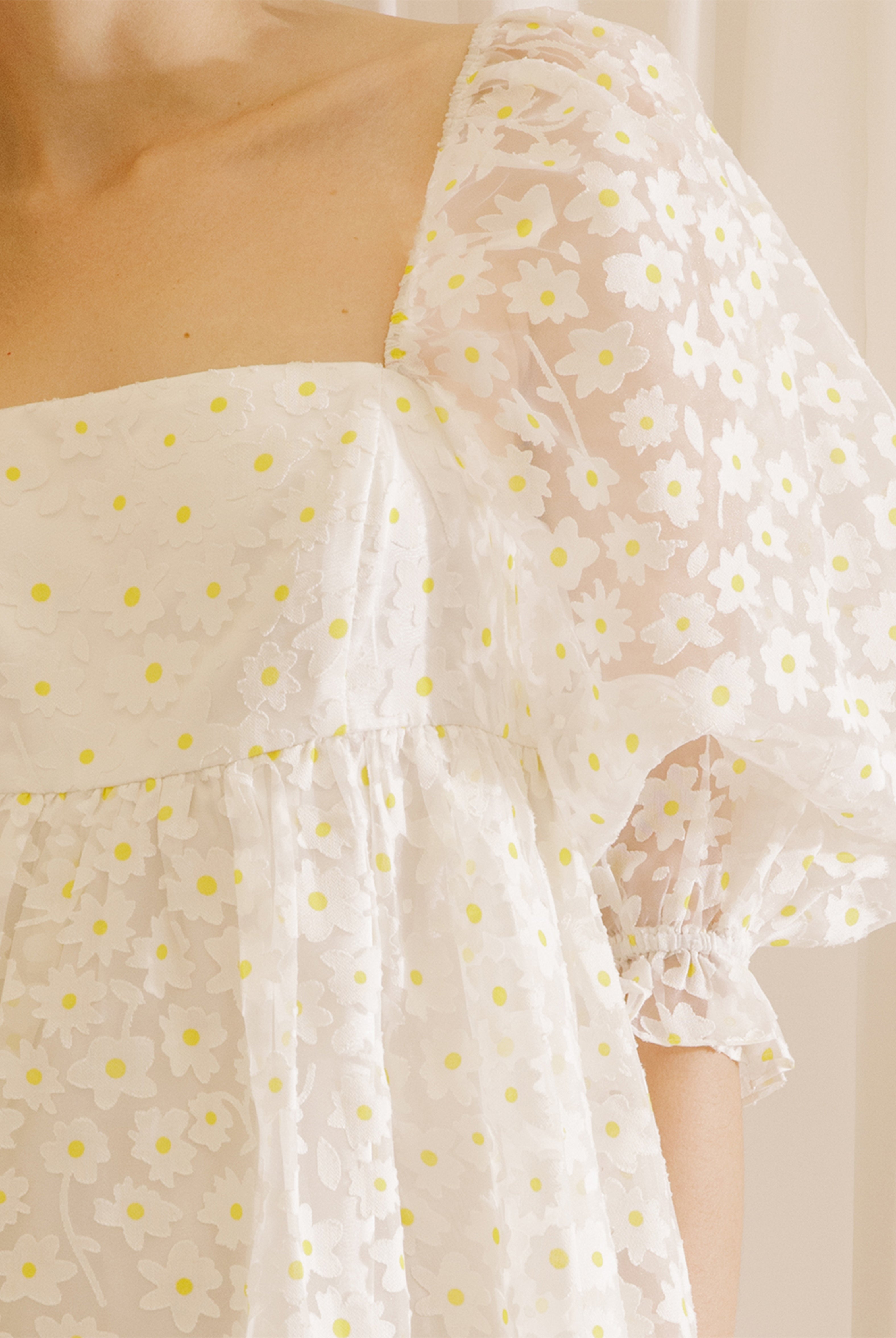 White dress with a sheer daisy-flocked fabric with yellow accents against a beige curtain background
