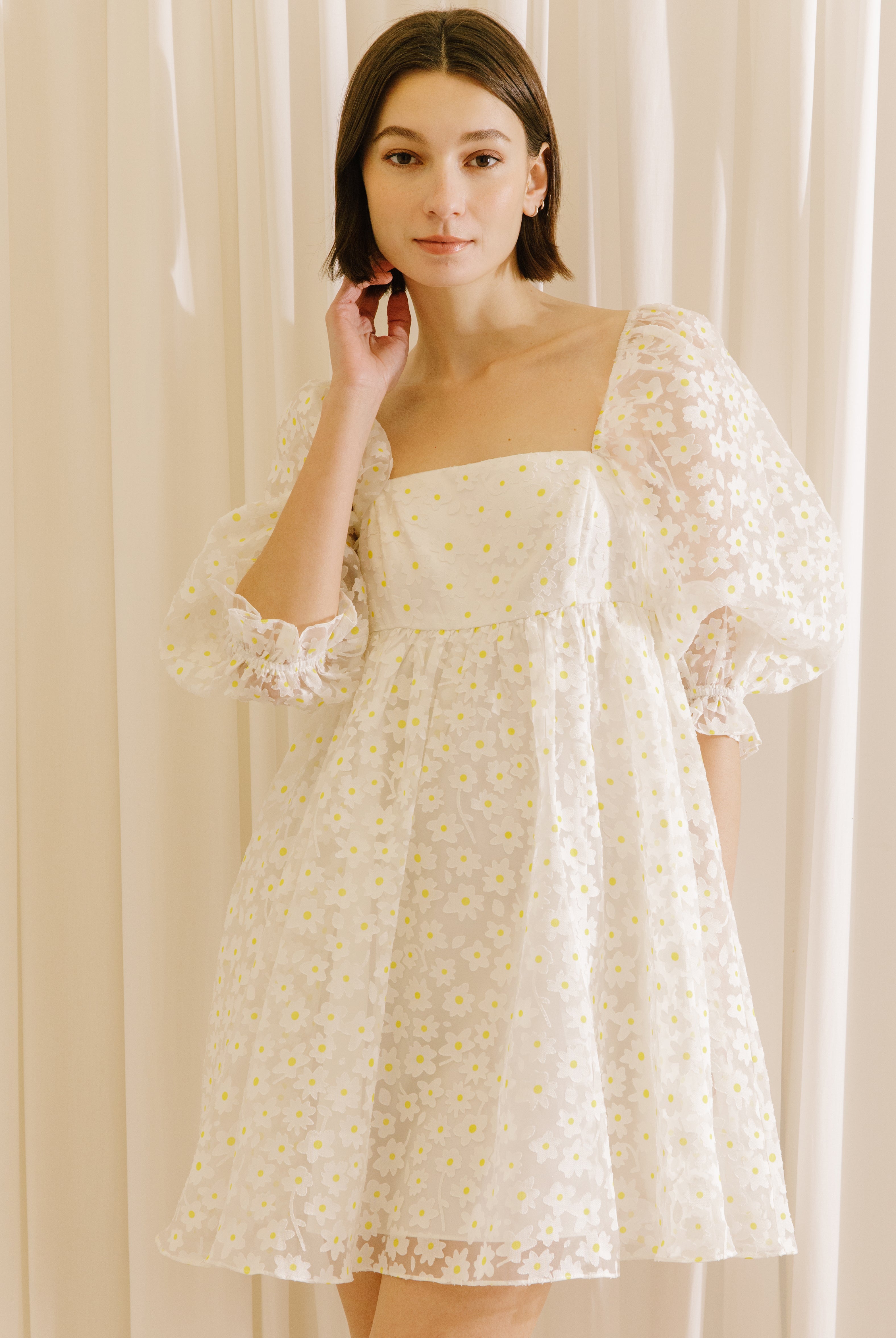 Woman wearing a white dress with a sheer daisy-flocked fabric with yellow accents against a beige curtain background
