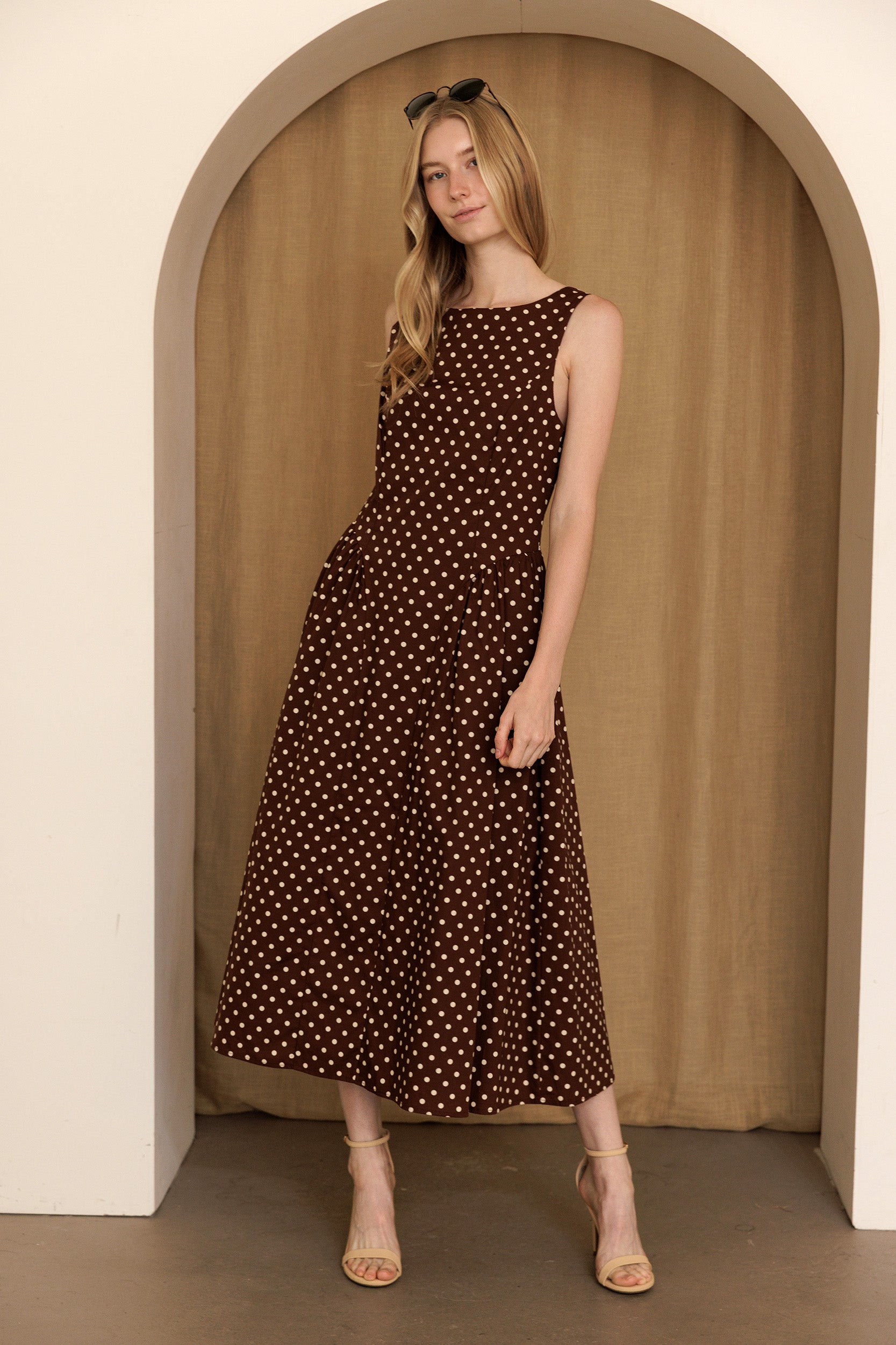 Woman wearing a brown polka dot midi dress standing in an archway with a beige curtain.