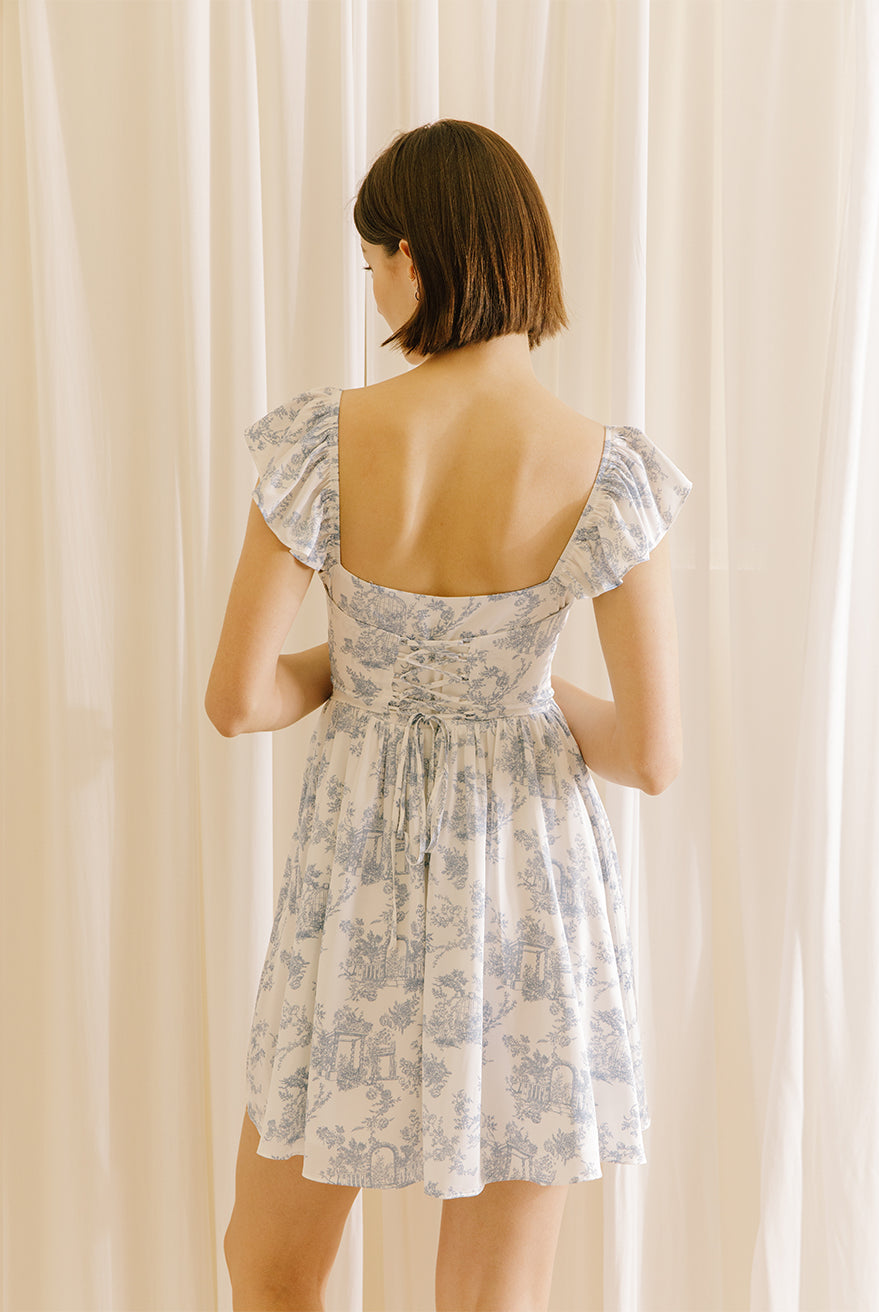 Back view of a woman wearing a white and blue floral mini dress with a corset crisscross back against a beige curtain background