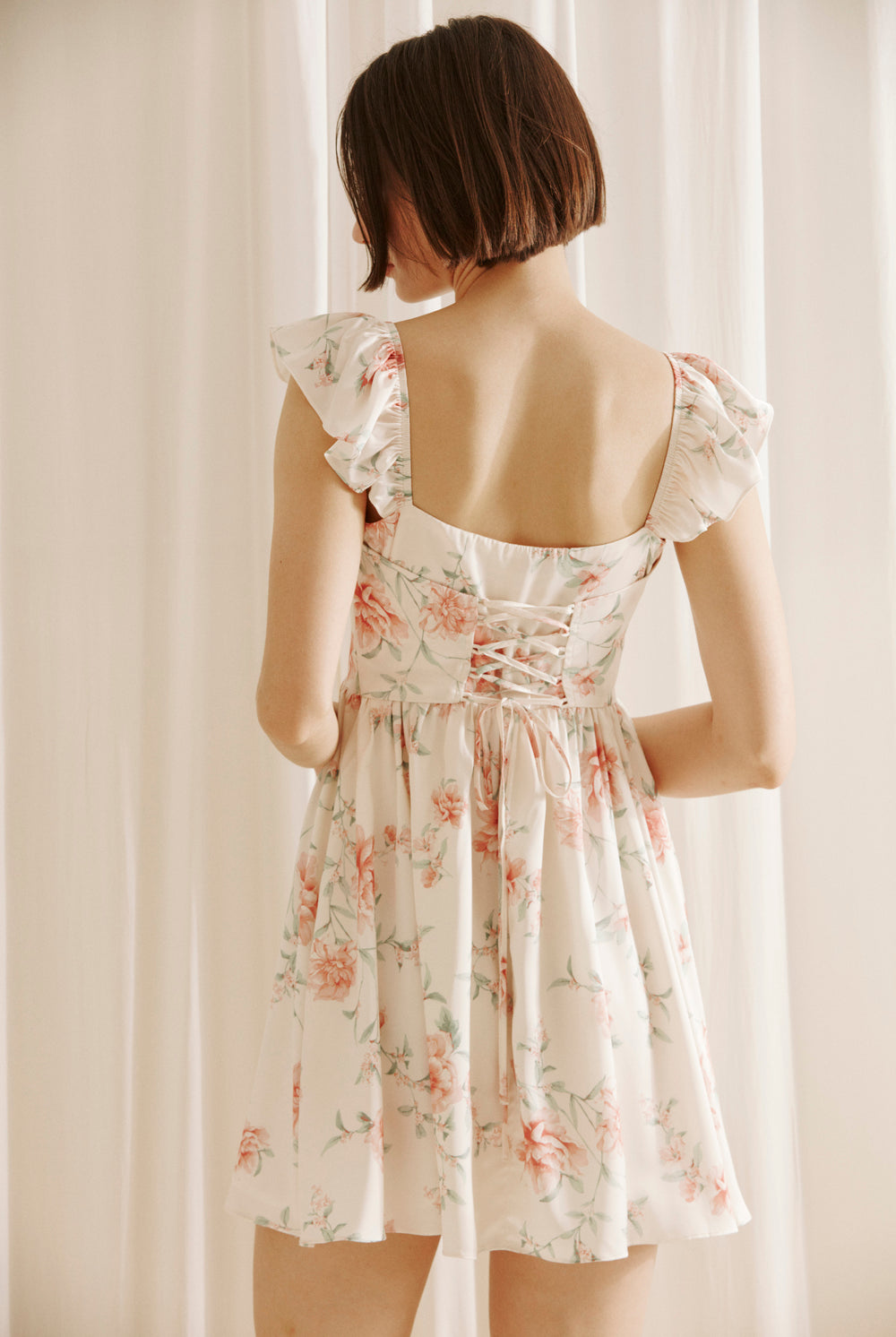 Back view of a woman wearing a cream floral mini dress with a corset crisscross back against a plain background
