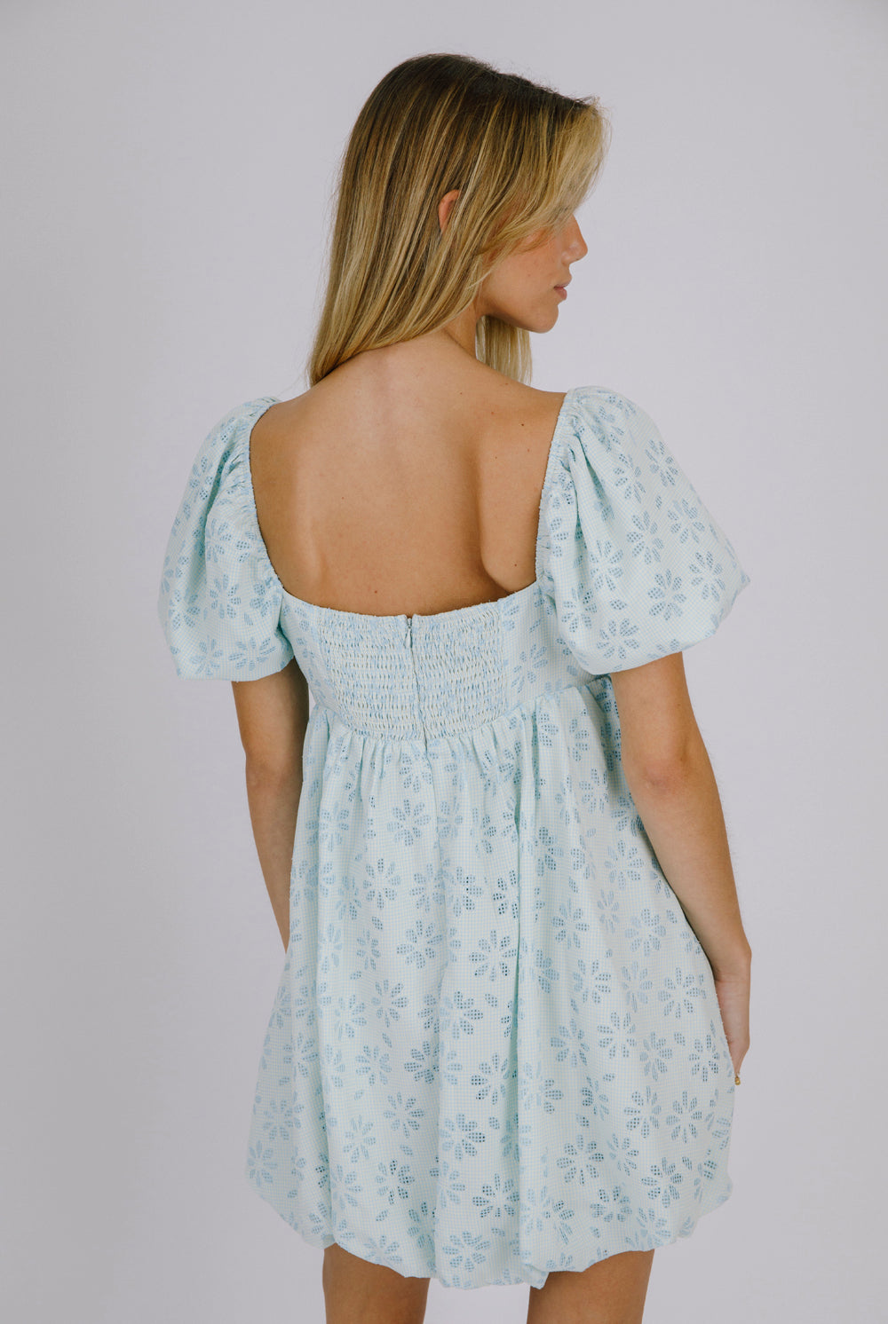 Back view of a woman wearing a blue gingham with floral embroidery mini dress against a plain background