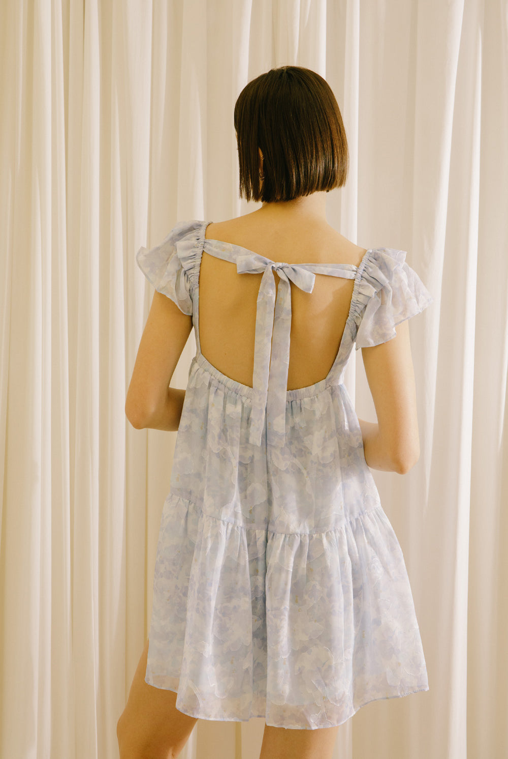 Back view of a woman wearing a light blue dress with a back self-tie bow against a beige curtain background