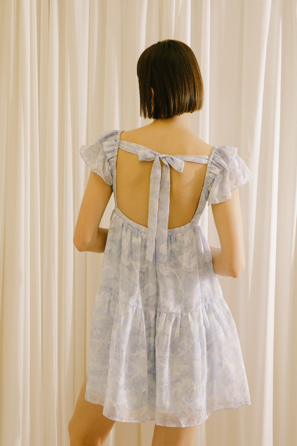 Back view of a woman wearing a light blue dress with a back self-tie bow against a beige curtain background