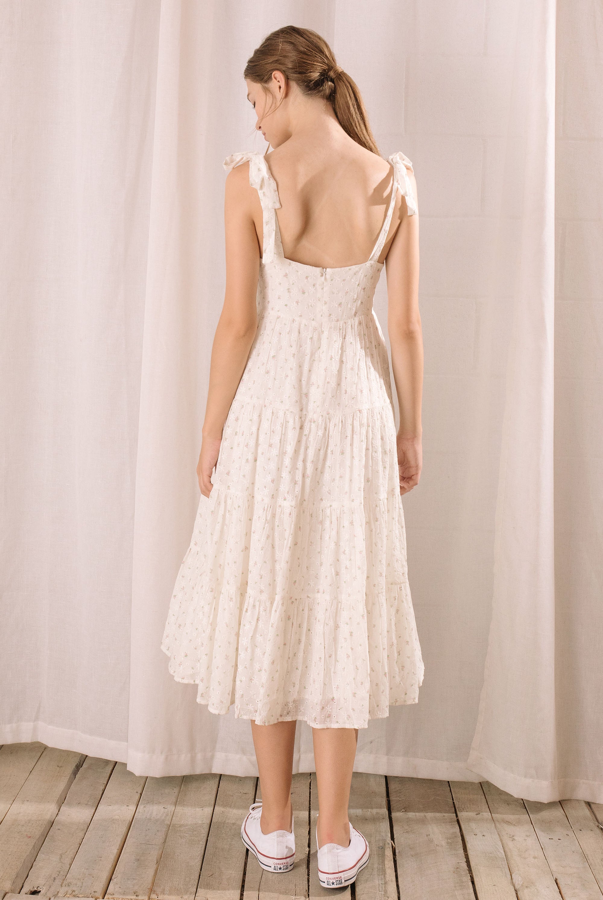 Back view of a woman wearing a white floral dress standing against a plain background
