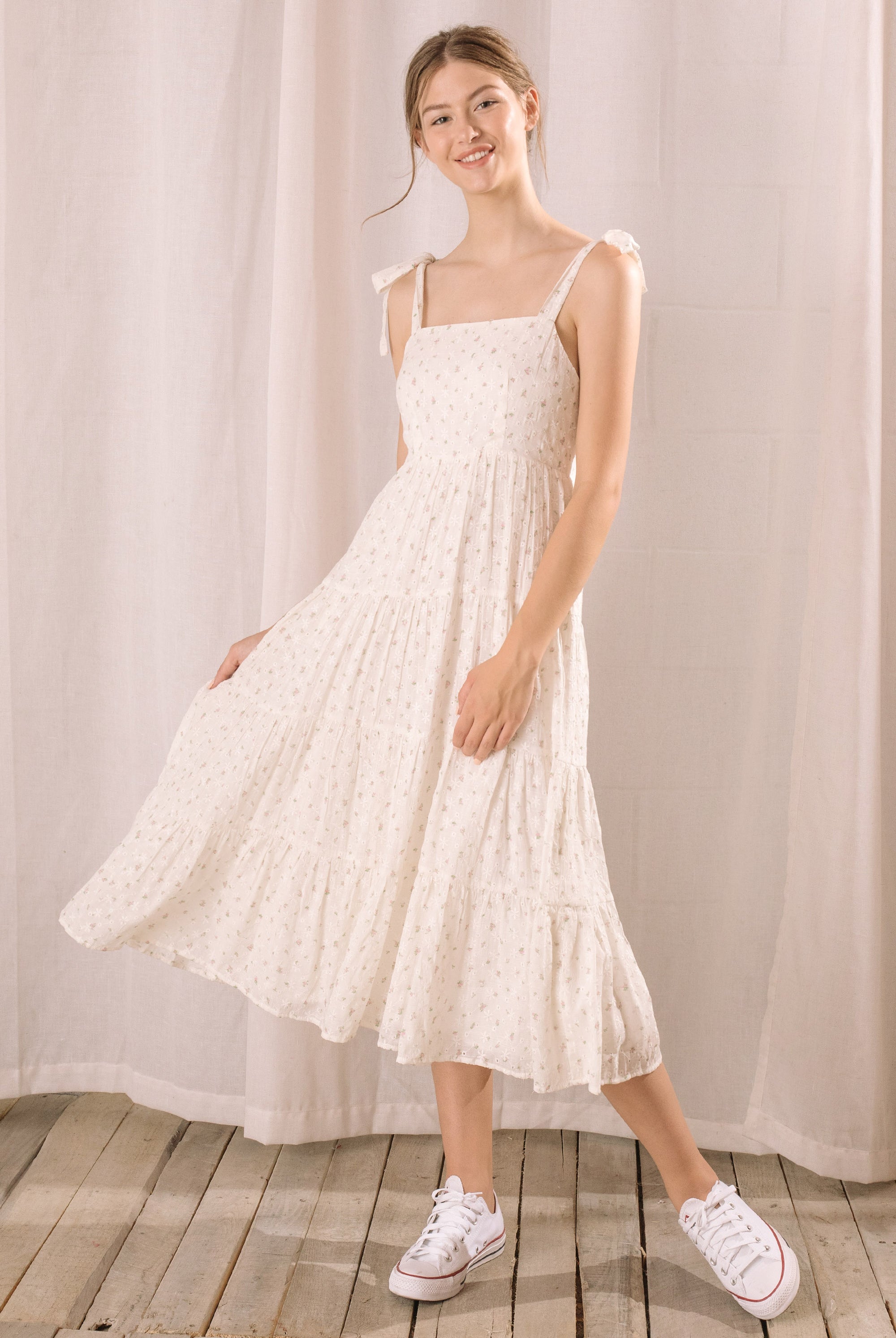 Woman wearing a white floral dress with white sneakers on a wooden floor and white curtain background