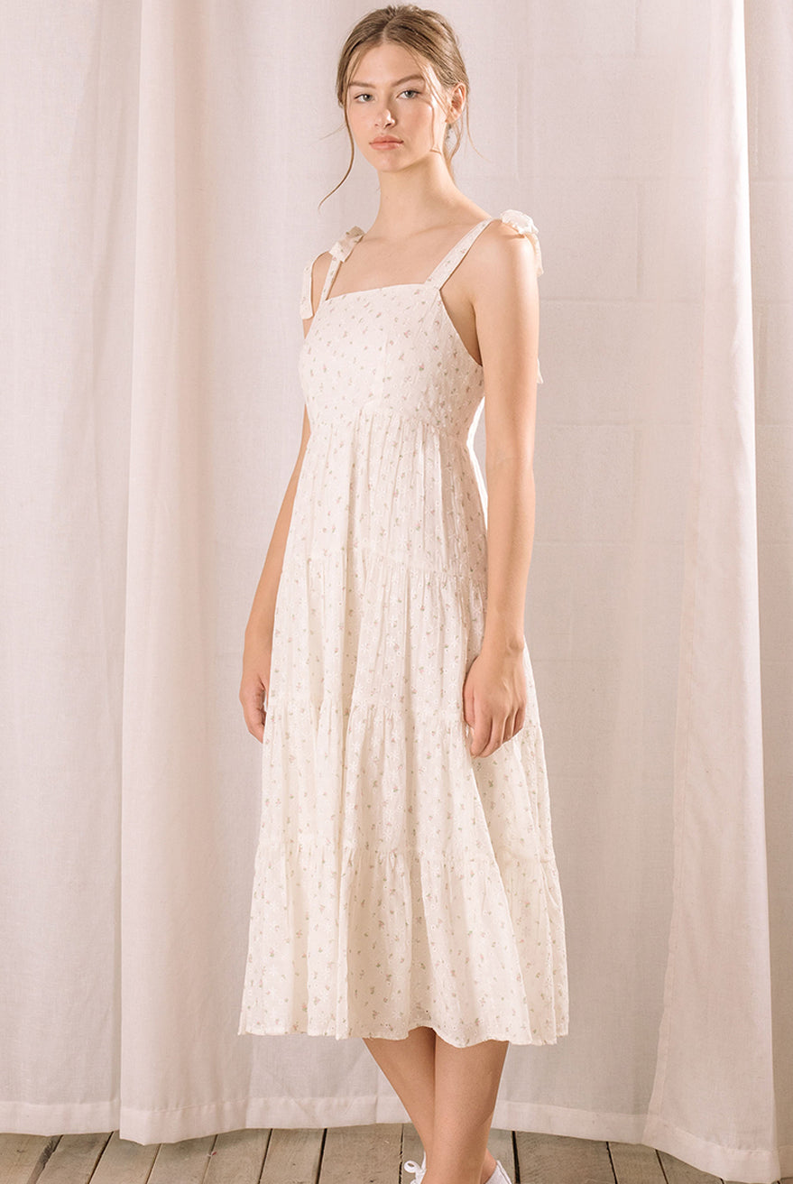 Side view of a woman wearing a white floral dress standing against a light curtain background