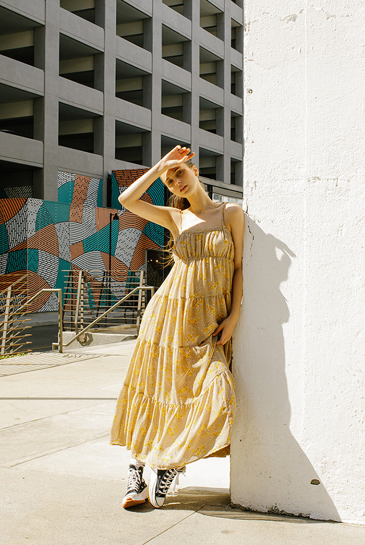 A woman standing against a white wall wearing a yellow floral ruffled maxi dress with a straight neckline, empire waist, and flowy skirt. She has her hand raised to her head.