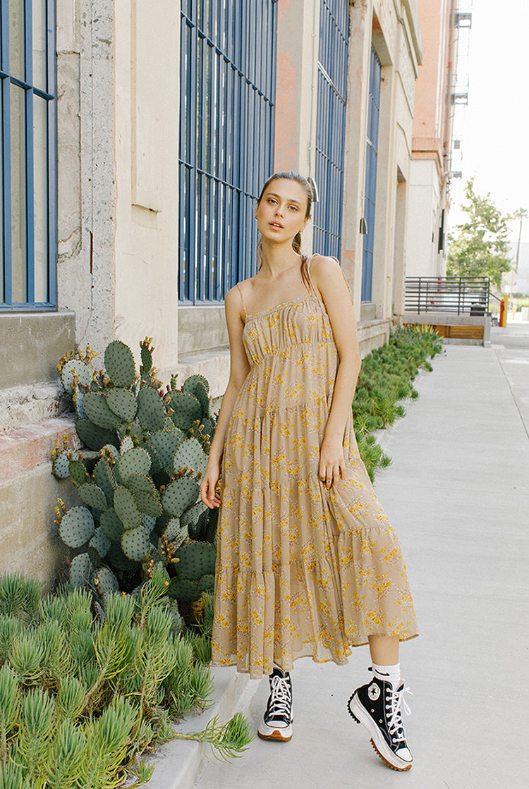A woman standing against a building wearing a yellow floral ruffled maxi dress with a straight neckline, empire waist, and flowy skirt.