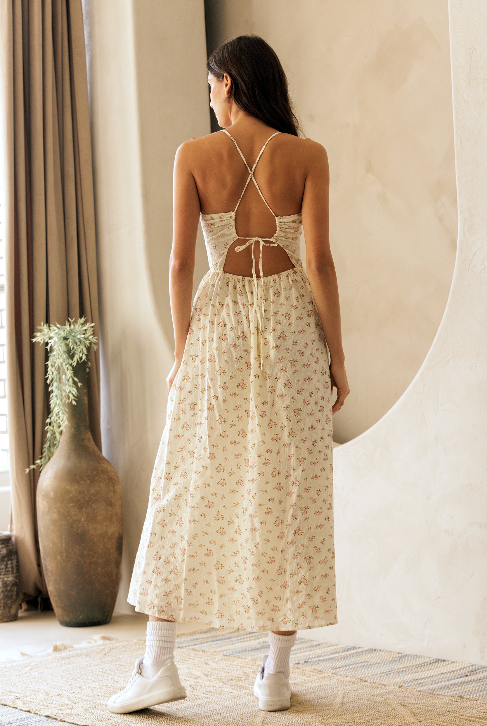 Back of a model wearing a floral dress with an open back in a room with beige walls and a vase.