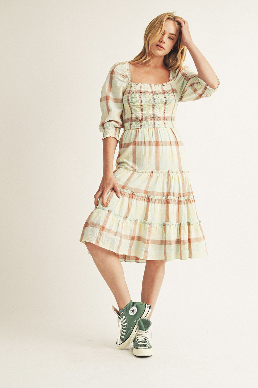 A woman modeling the Elliana Mint Large Plaid Midi Dress with a square neckline, puffy sleeves, and a smocked upper body. The dress features a neutral and pastel plaid pattern and she is wearing green sneakers.