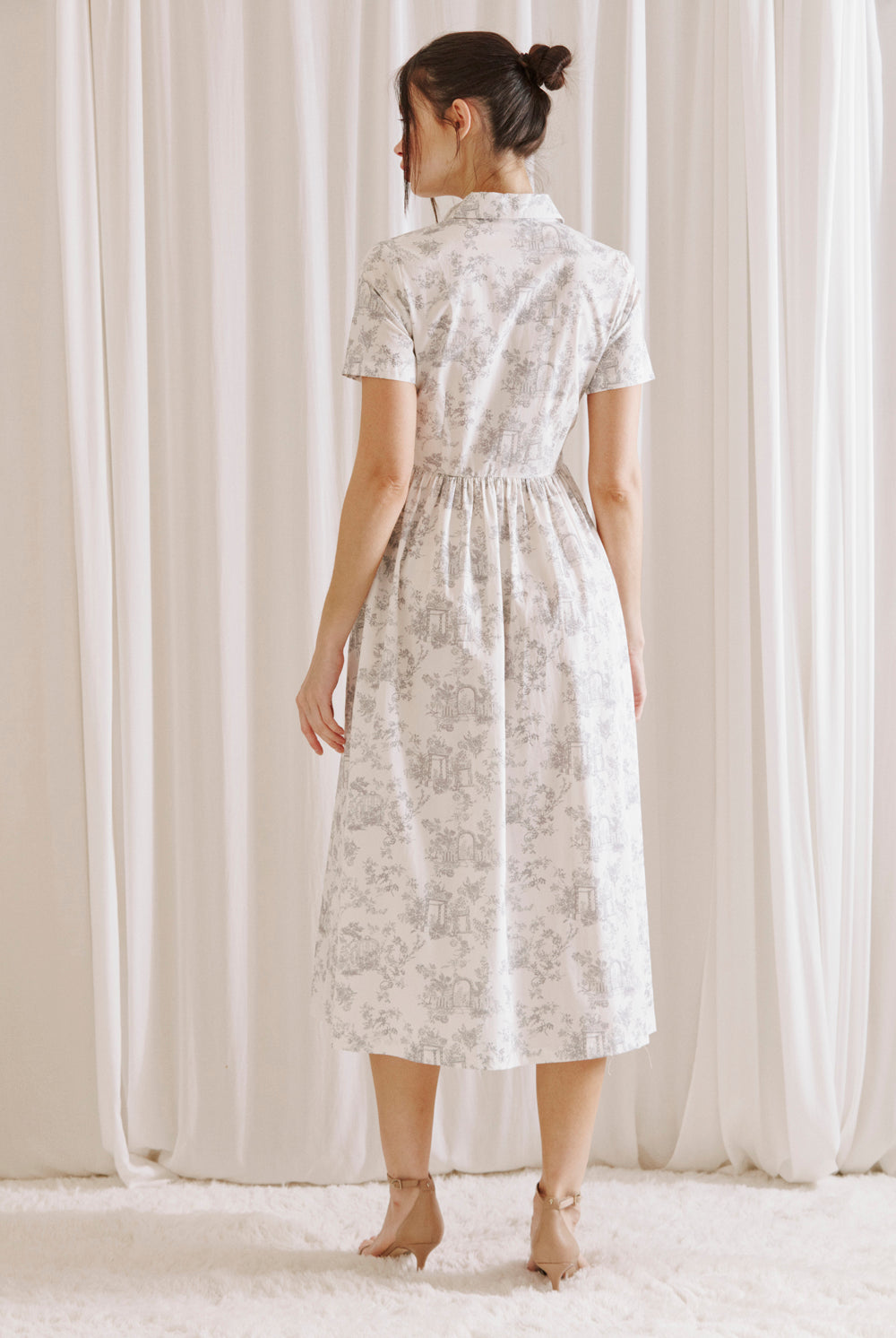 Back view of a woman wearing a white and gray toile print midi dress standing against a white curtain backdrop