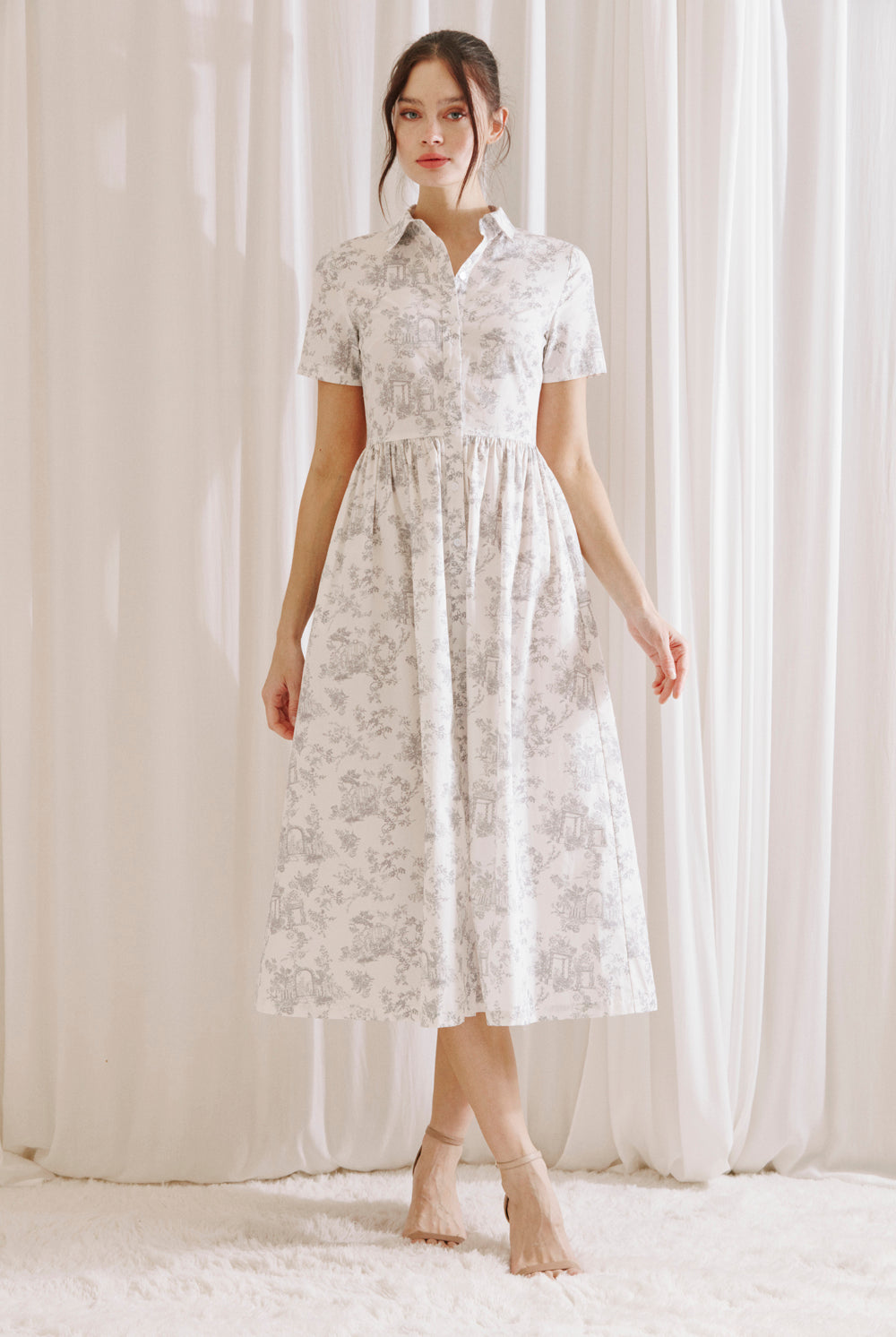 Woman wearing a white and gray toile print midi dress standing against a white curtain backdrop