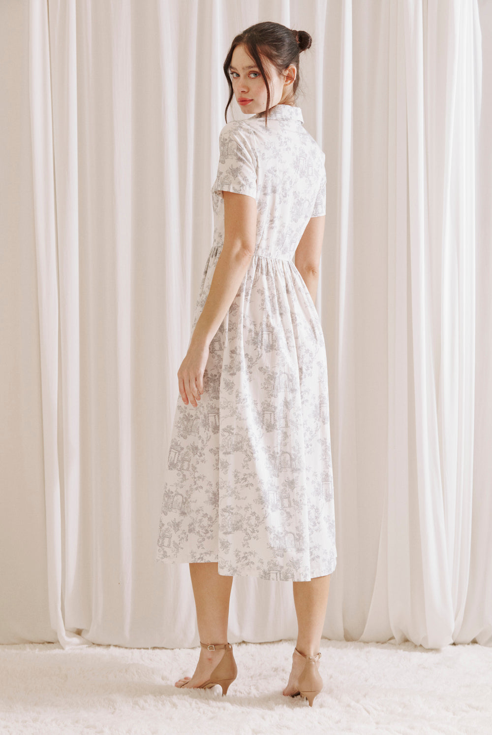 Side view of a woman wearing a white and gray toile print midi dress standing against a white curtain backdrop