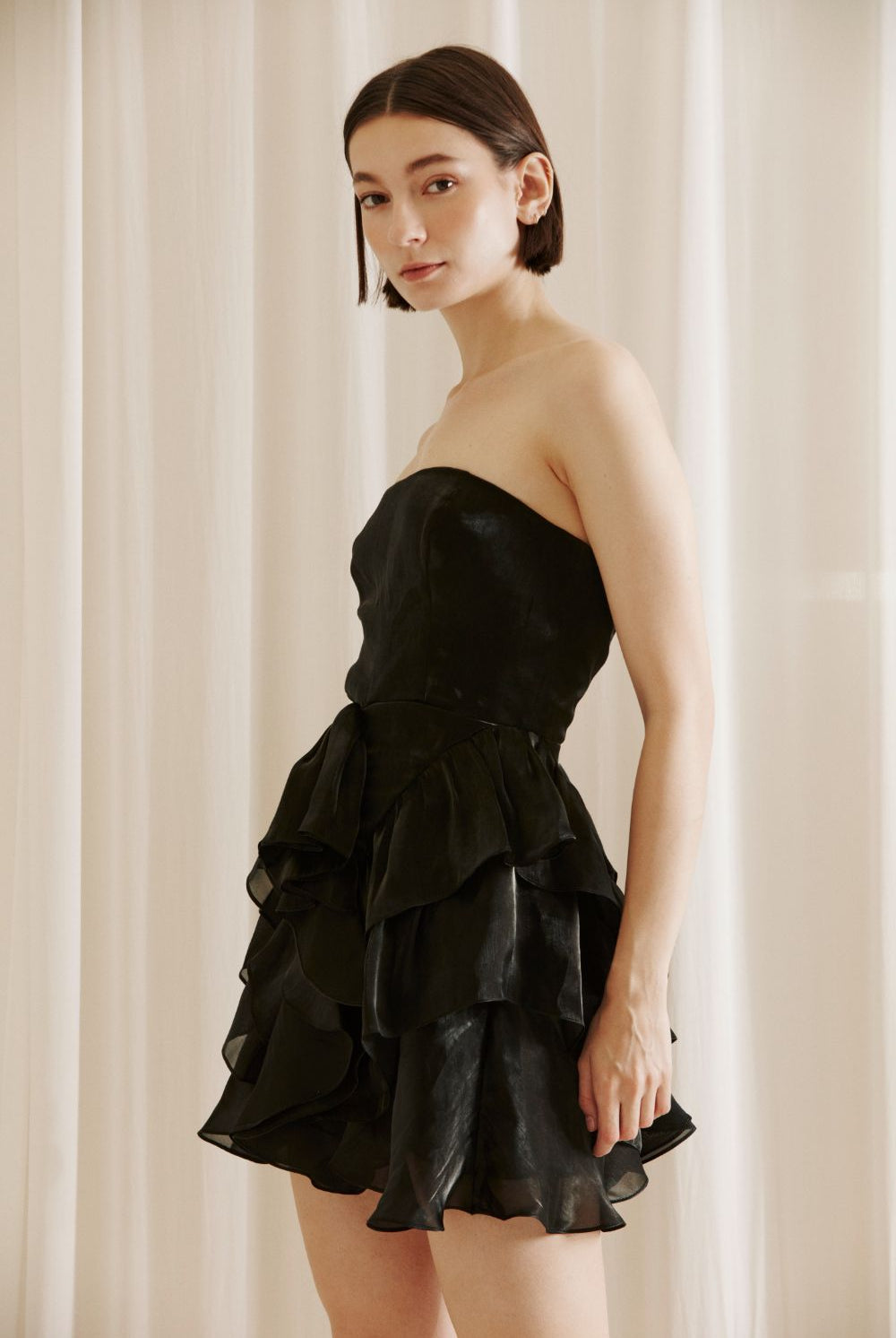 Side view of a woman wearing a black strapless dress with ruffled layers against a beige curtain background