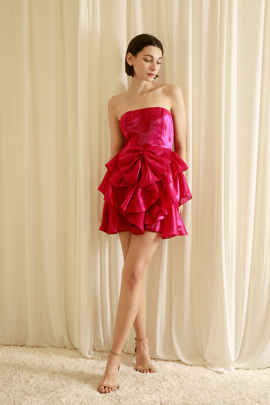 Woman wearing a bright pink strapless dress with ruffled details and beige heels against a beige wall.