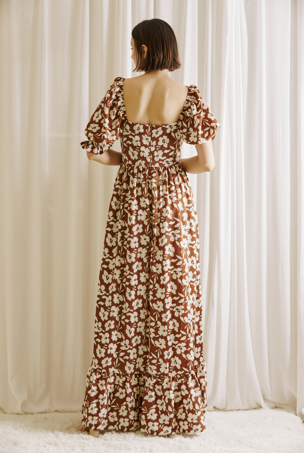 Back view of a woman wearing a brown and white floral dress standing against a white curtain backdrop