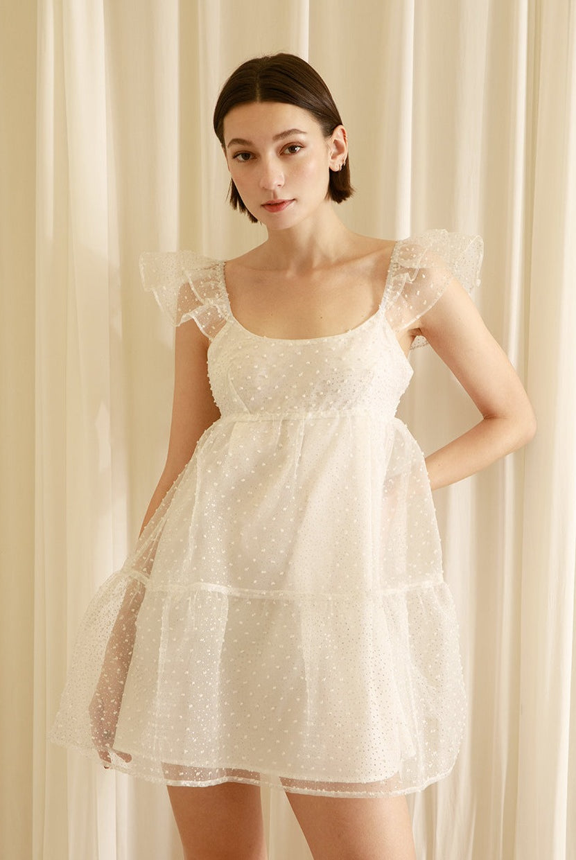 Woman wearing a white swiss dot overlay dress against a beige curtain background