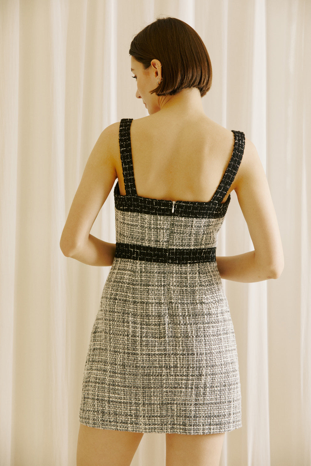 Back view of a woman wearing a black and white checkered tweed dress with an invisible back zipper against a white curtain background