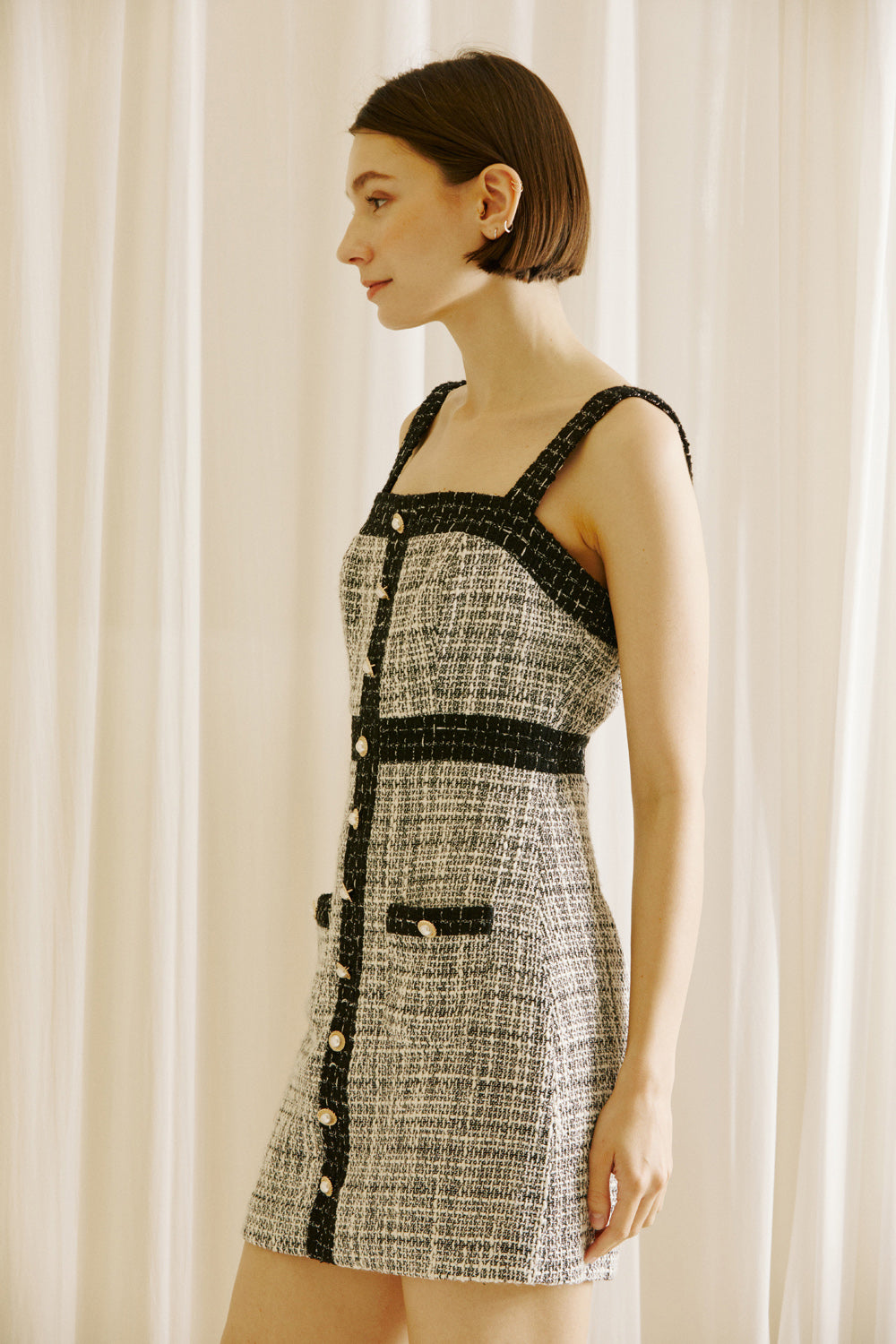 Side view of a woman wearing a black and white checkered tweed dress with pearl button details against a white curtain background