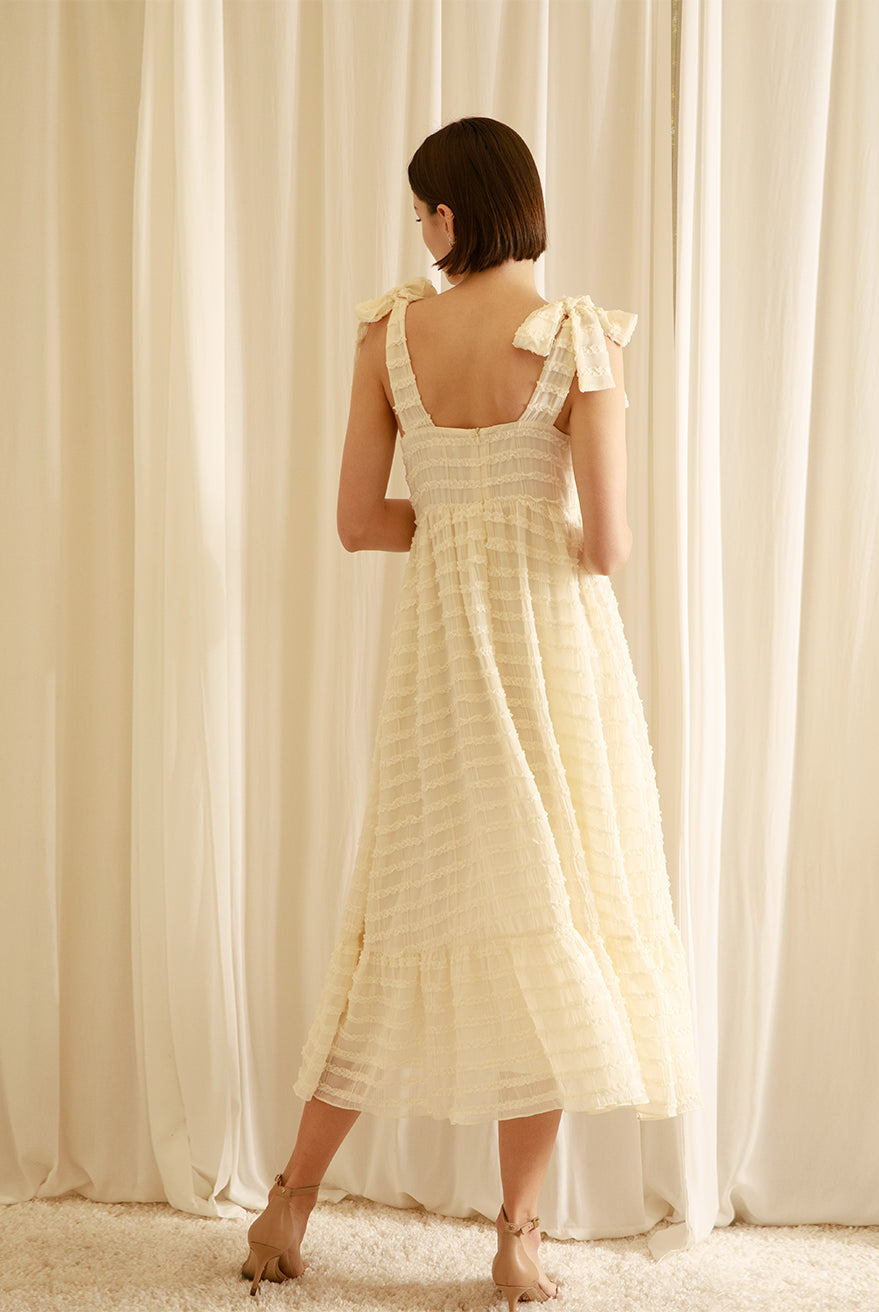 Back view of a woman wearing a cream midi dress standing in front of white curtains