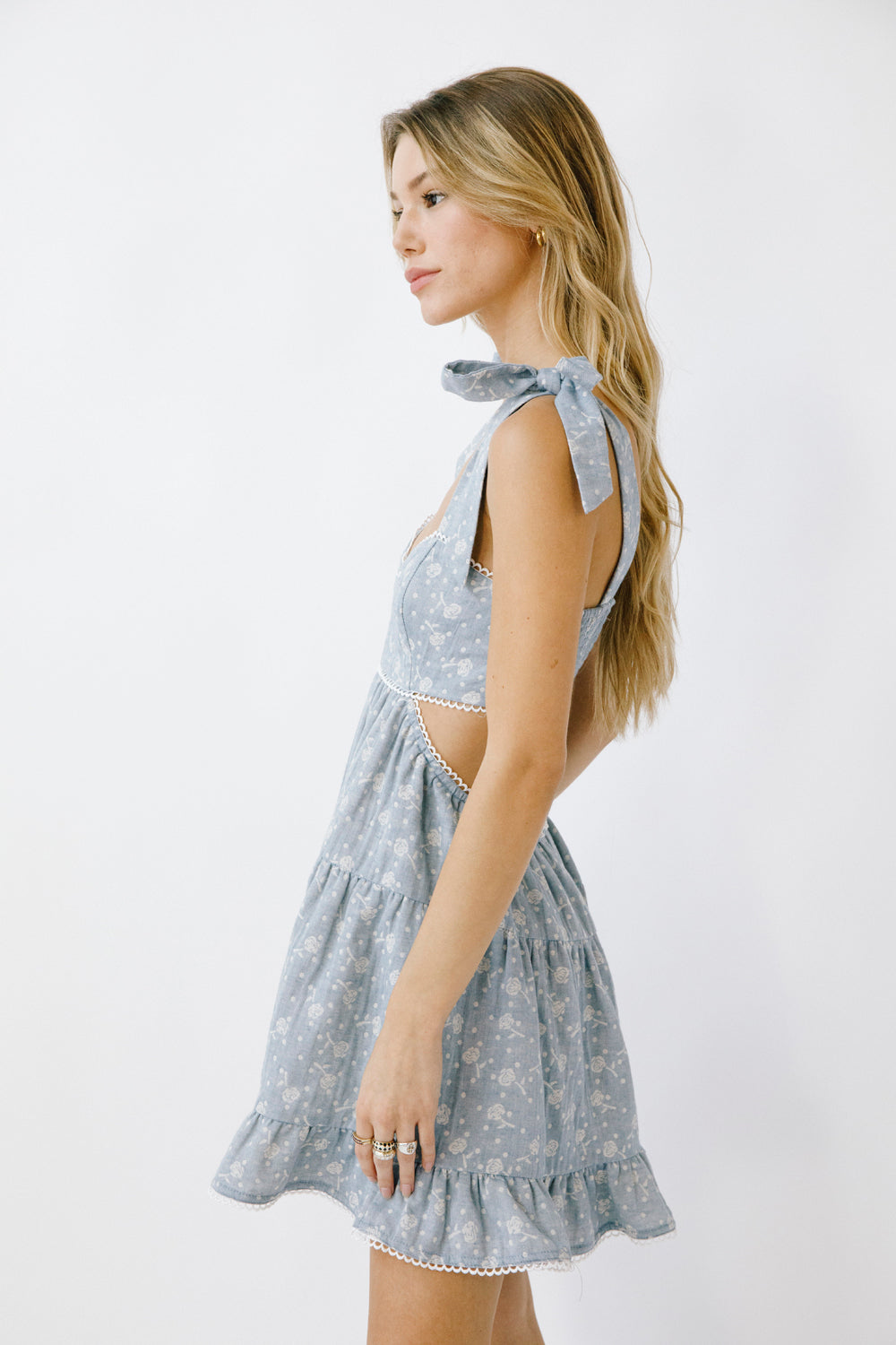 A woman wearing a blue denim mini dress with a rose print, adjustable straps, and a ruffled hem.