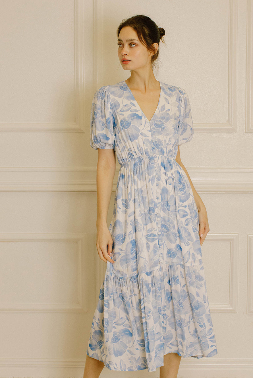 A woman wearing a light blue and white floral midi dress with puff sleeves and a V-neckline, standing against a cream-colored paneled wall.
