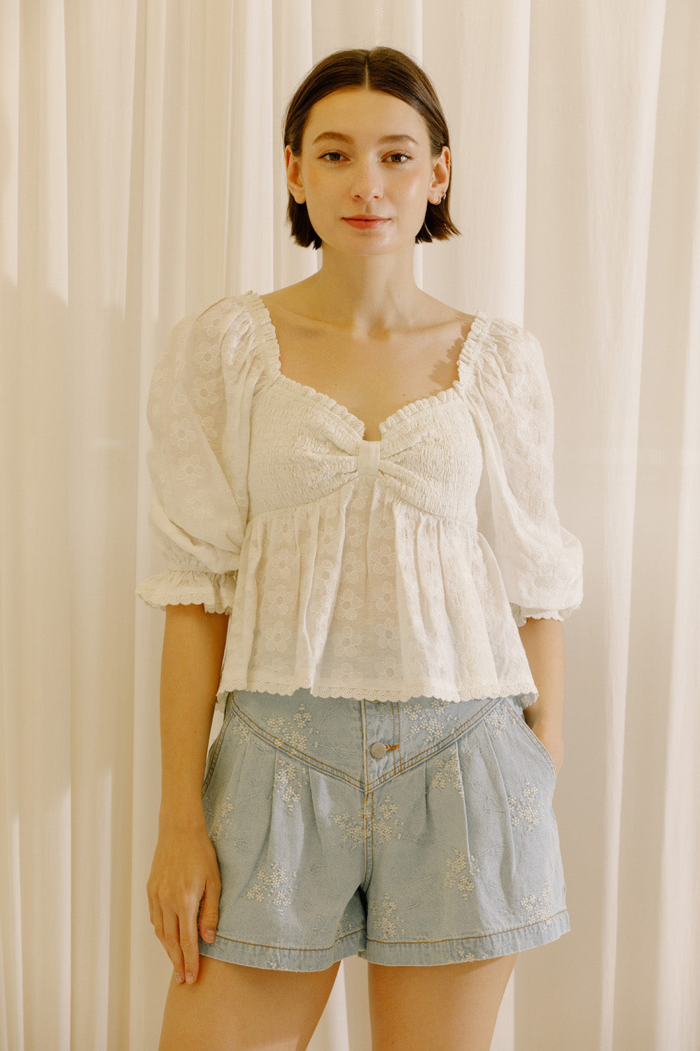 A woman wearing a white embroidered puff-sleeve blouse with a sweetheart neckline and a knotted front detail, paired with light blue denim shorts, standing in front of a cream-colored curtain backdrop.