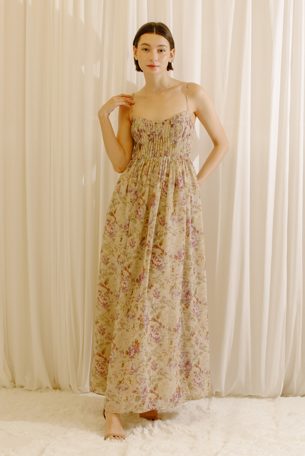A model wearing a maxi dress with a romantic floral print, pleated bustier bodice, sweetheart neckline, adjustable straps, and a flowy maxi silhouette.