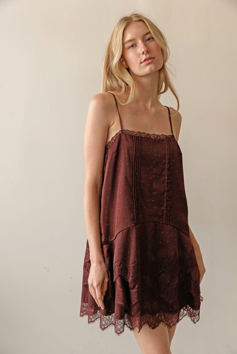 A blonde woman wearing a dark brown spaghetti-strap mini dress with lace trim, standing against a plain beige wall.