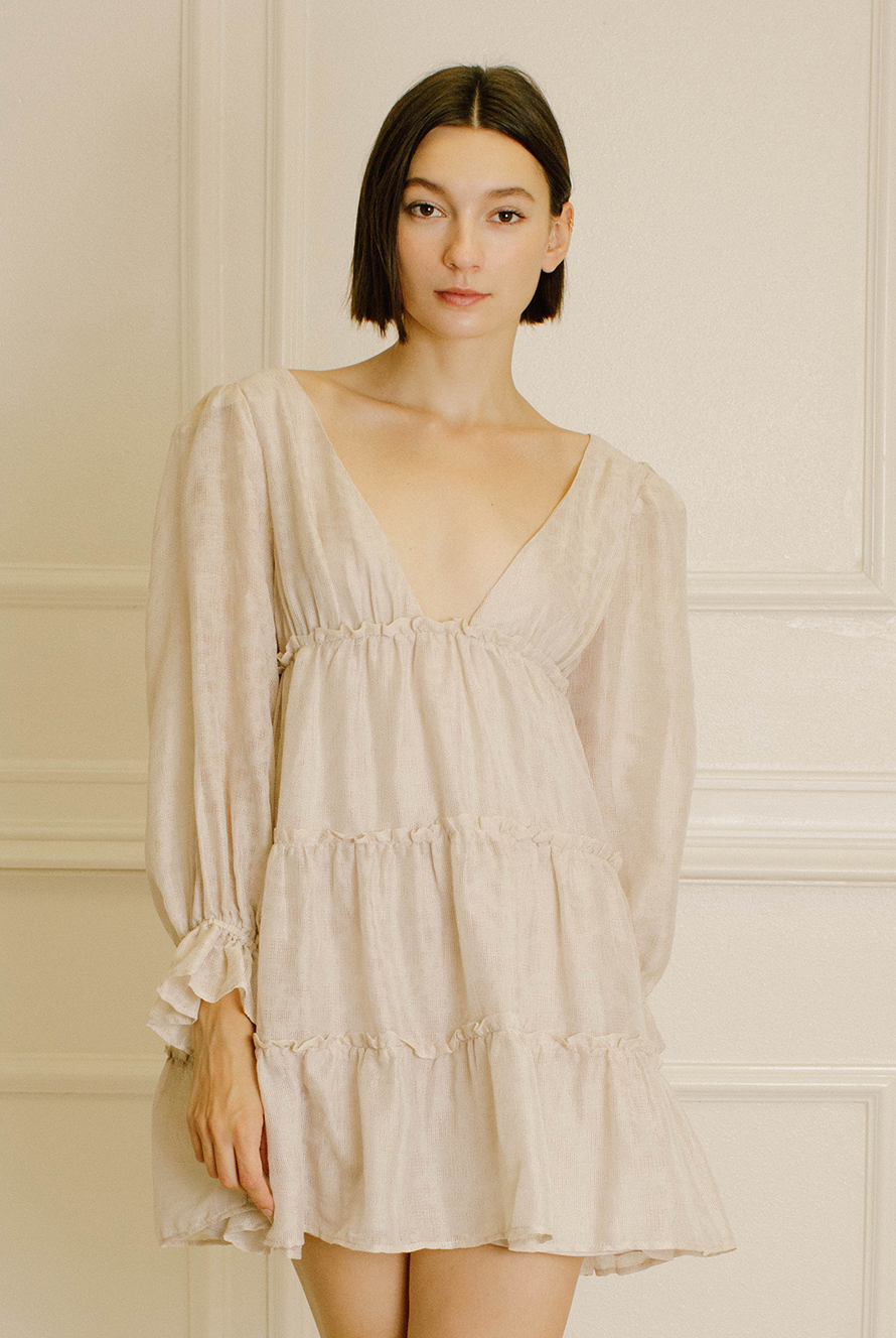 A woman with short dark hair wearing a light beige tiered mini dress with long sleeves, standing against a cream-colored paneled wall.