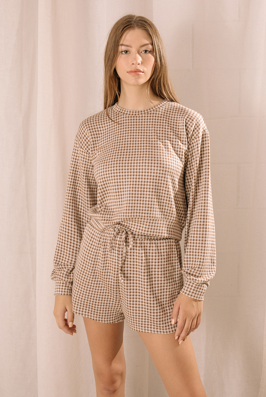 A woman modeling a lounge set that features a relaxed long-sleeve top and high-waisted drawstring shorts in a neutral brown and ivory gingham print.