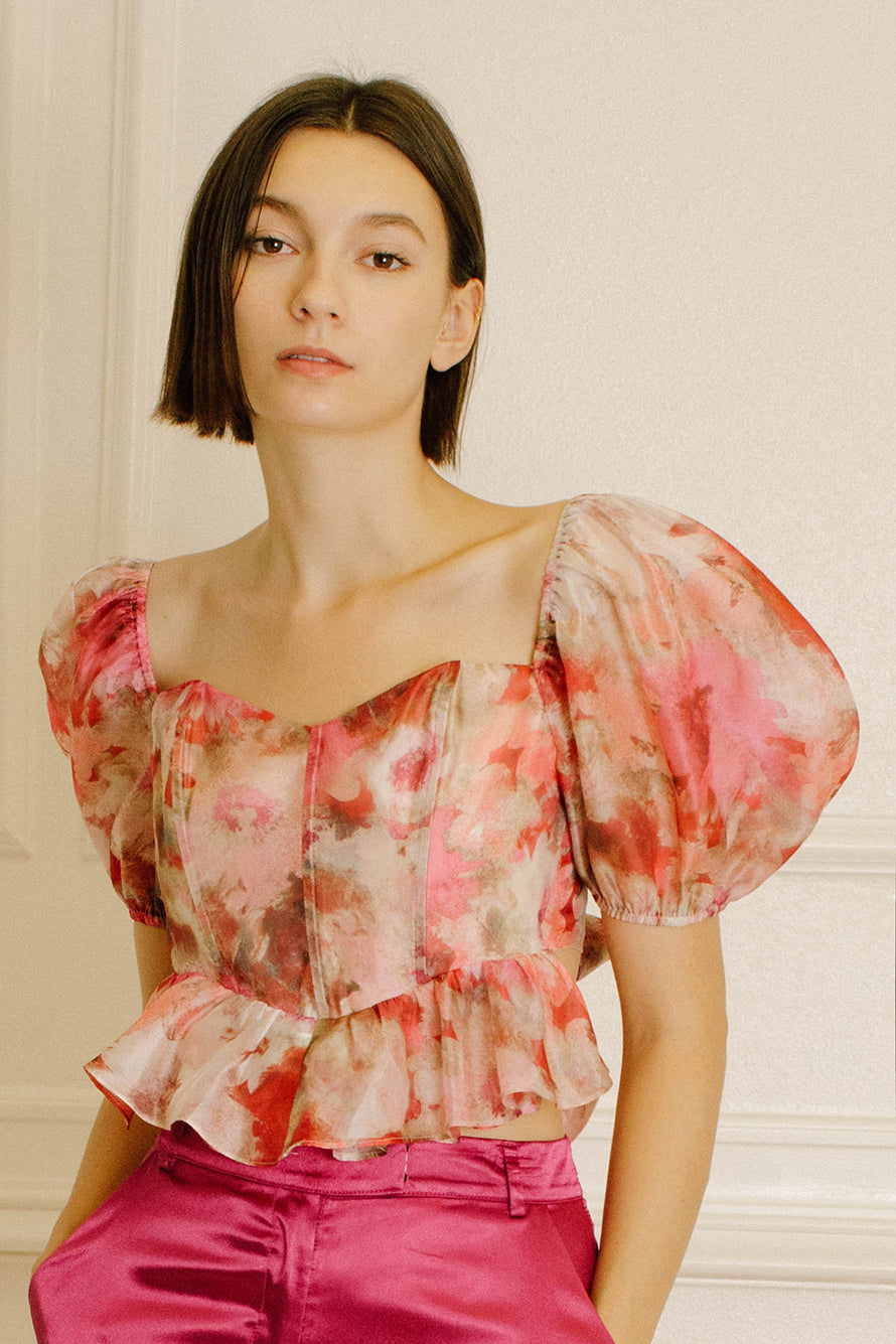 A woman wearing a sheer puff-sleeve blouse with a pink, red, and beige floral print, paired with bright pink satin pants, set against a light cream paneled wall.