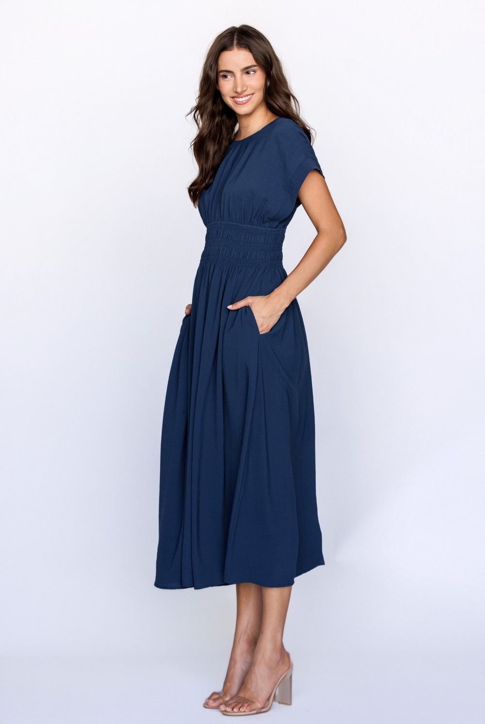 A model wearing a navy midi dress with a smocked waist, relaxed bodice, dolman sleeves, and side pockets against a white wall.