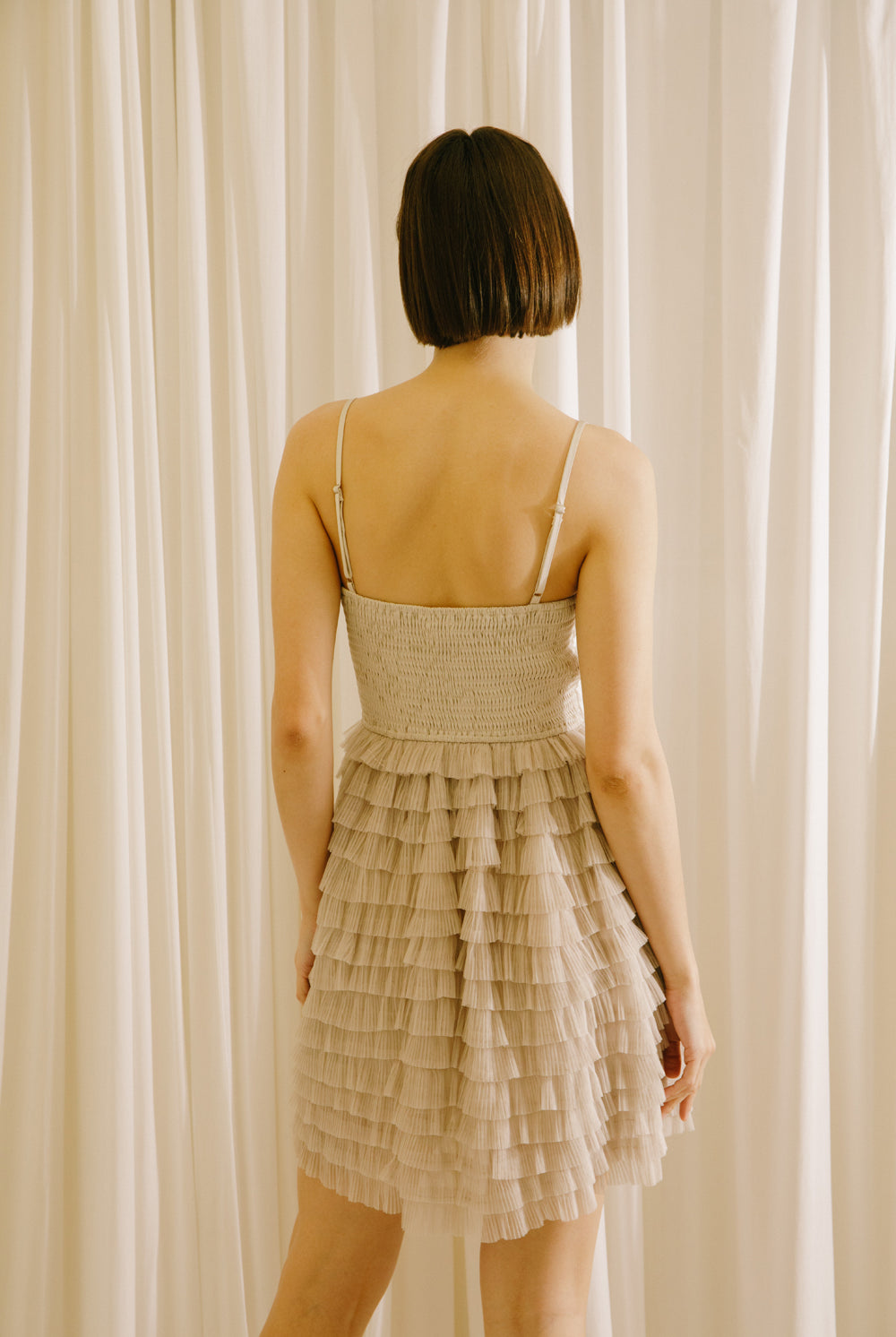 Back of a woman wearing a beige dress with a ruffled skirt against a white curtain background