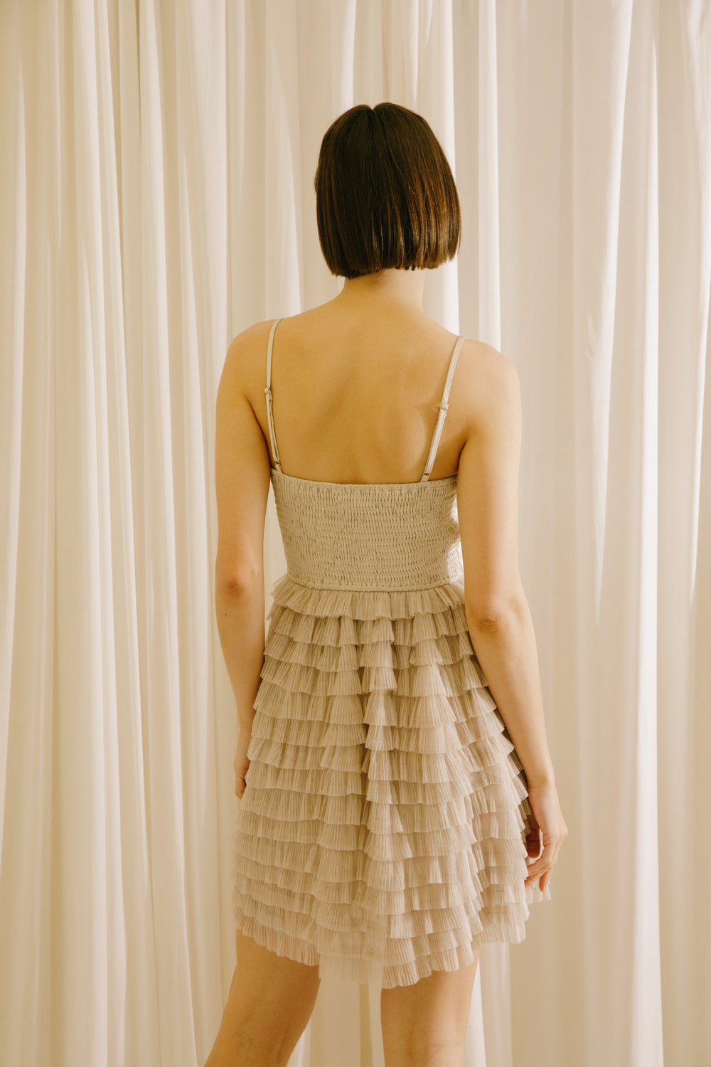 Back of a woman wearing a beige dress with a ruffled skirt against a white curtain background